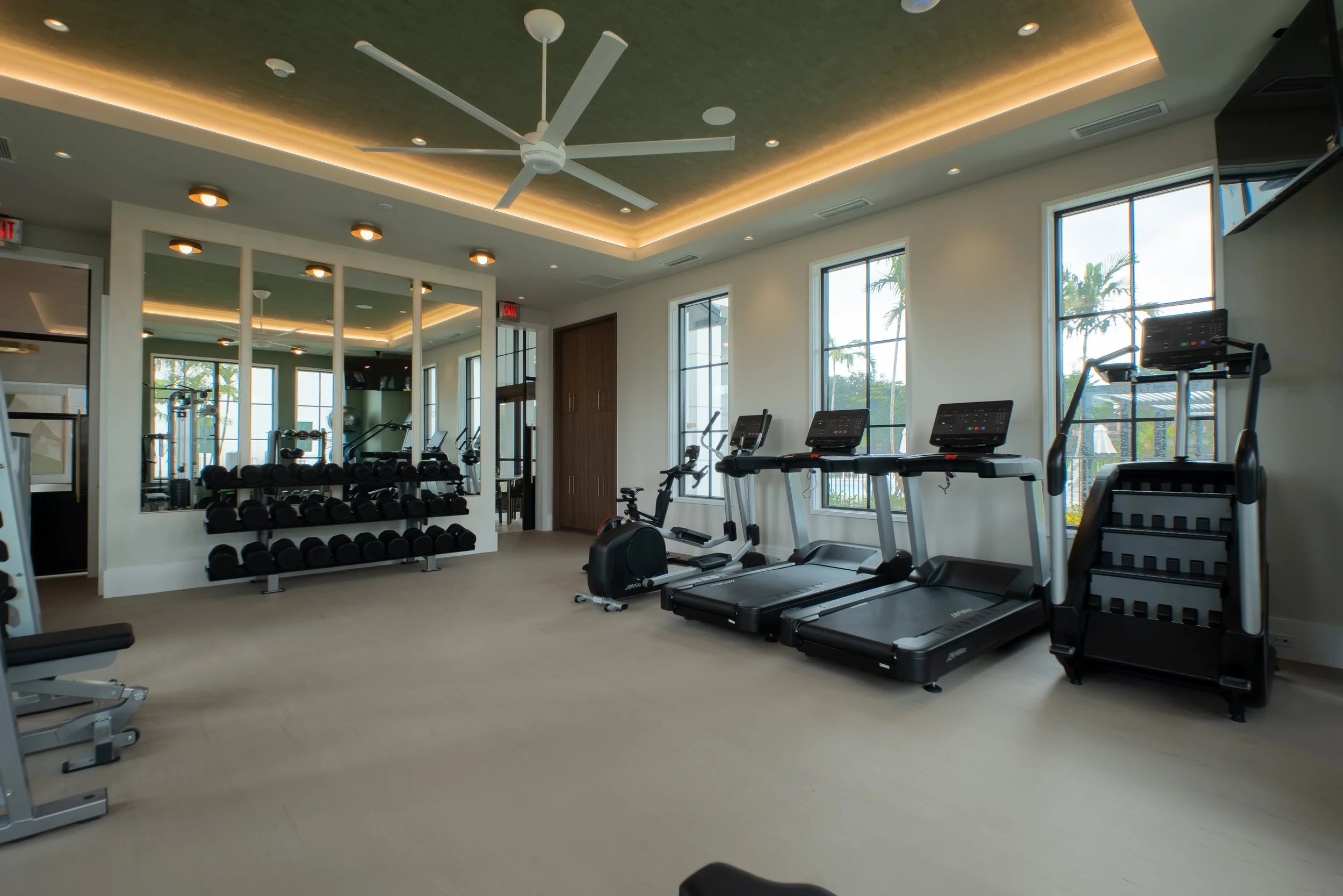 A modern gym room with exercise equipment, including treadmills and stationary bikes, large windows with a view of palm trees outside, and mirrors reflecting the ceiling with a ceiling fan and recessed lighting.