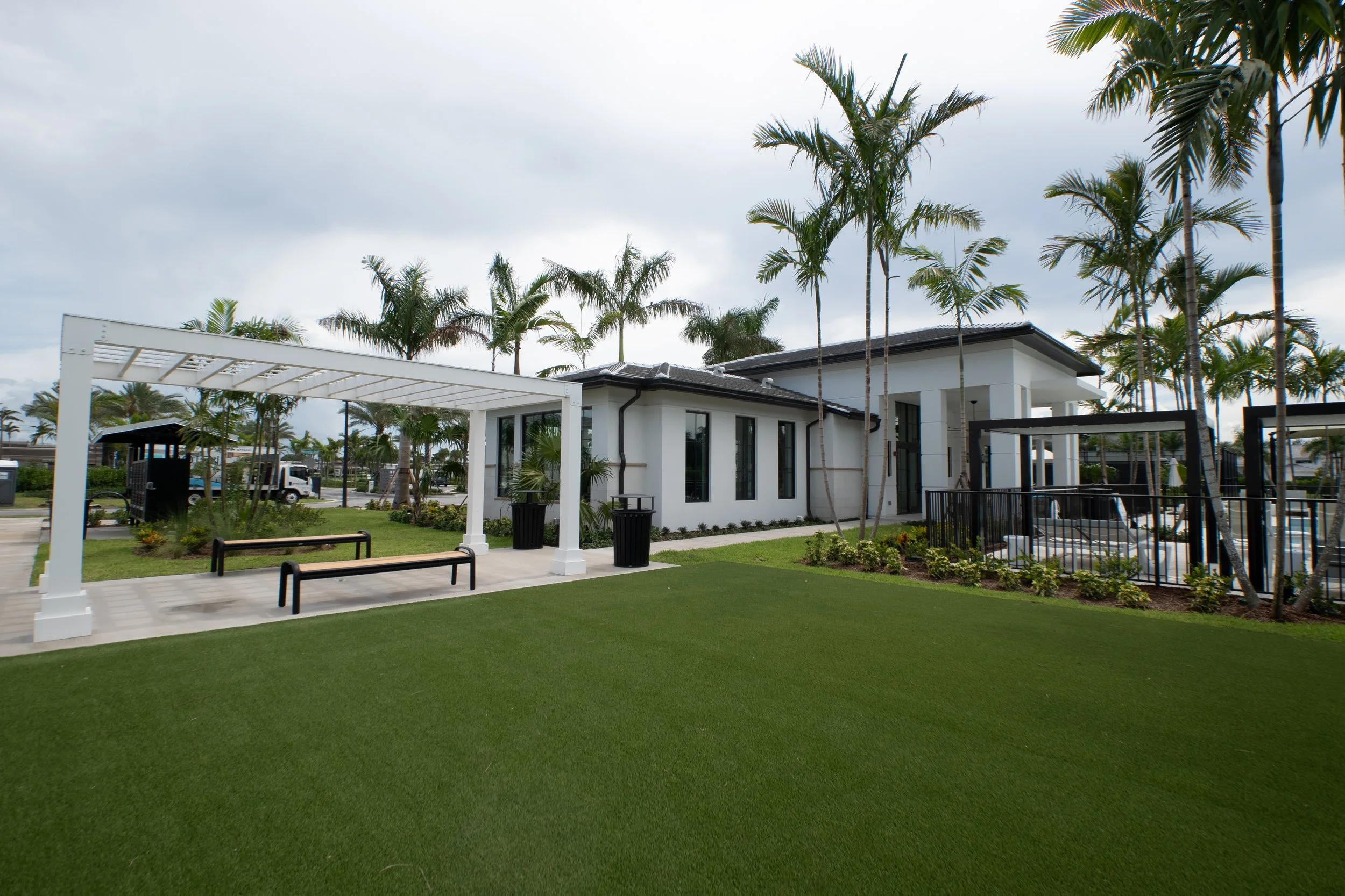 Outside area with green artificial grass, benches, white pergola, and modern white building with large windows, palm trees, and cloudy sky.