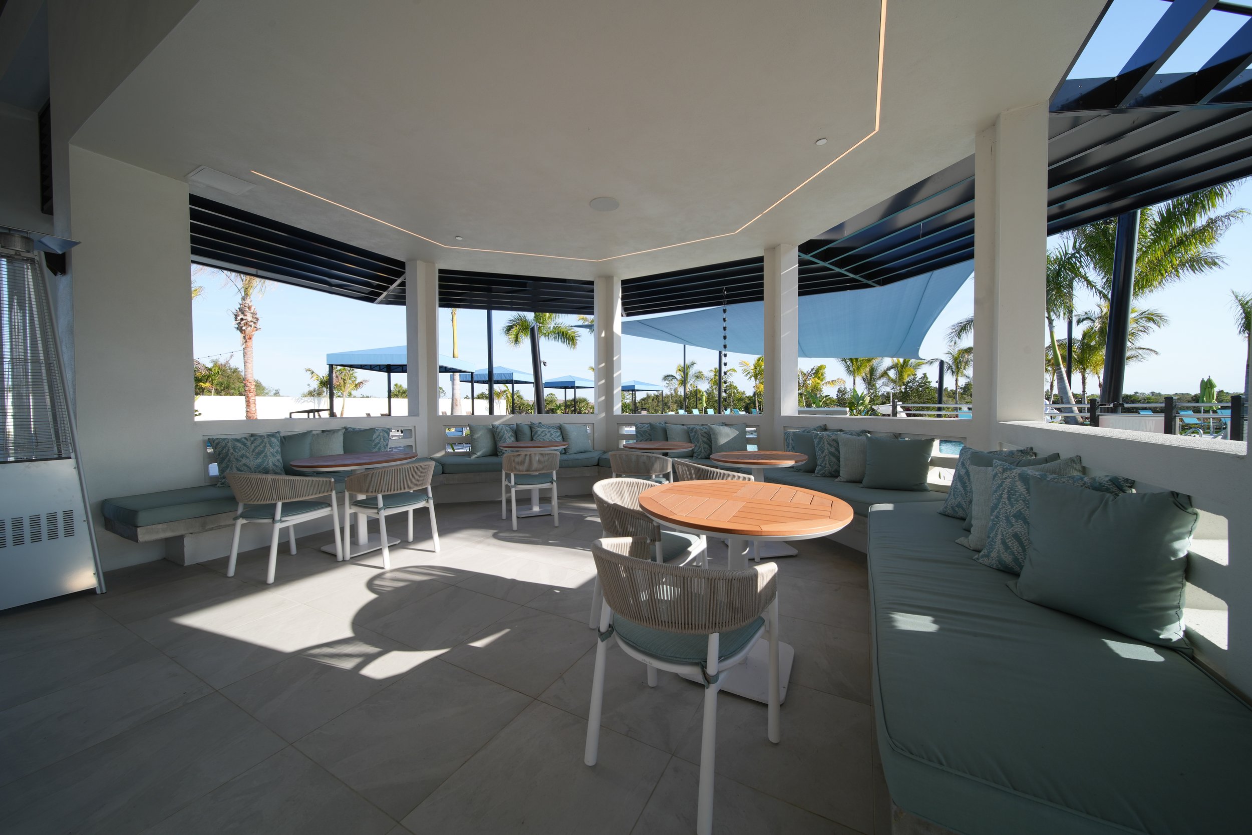 A covered outdoor lounge area with white cushioned seating, round wooden tables, and decorative pillows. Large windows reveal palm trees and blue sky outside.