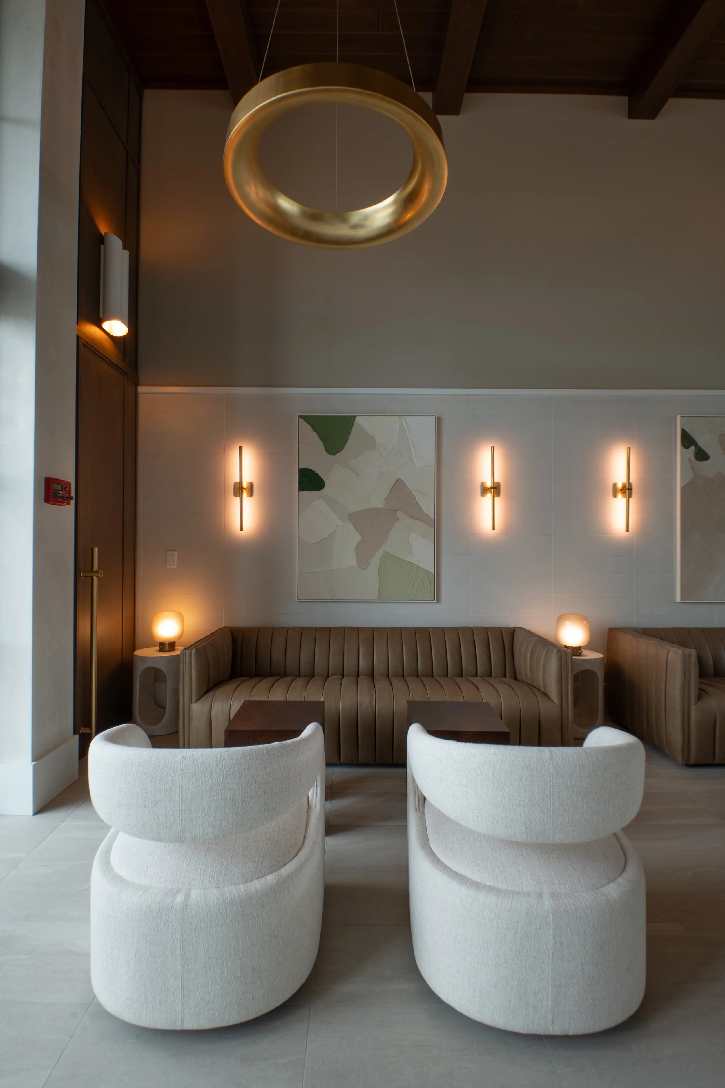 A modern interior with a pair of cream-colored lounge chairs in the foreground, a brown tufted sofa in the middle, a wooden coffee table, and minimalistic wall sconces and paintings on the back wall. A gold decorative ring pendant hangs from the ceil