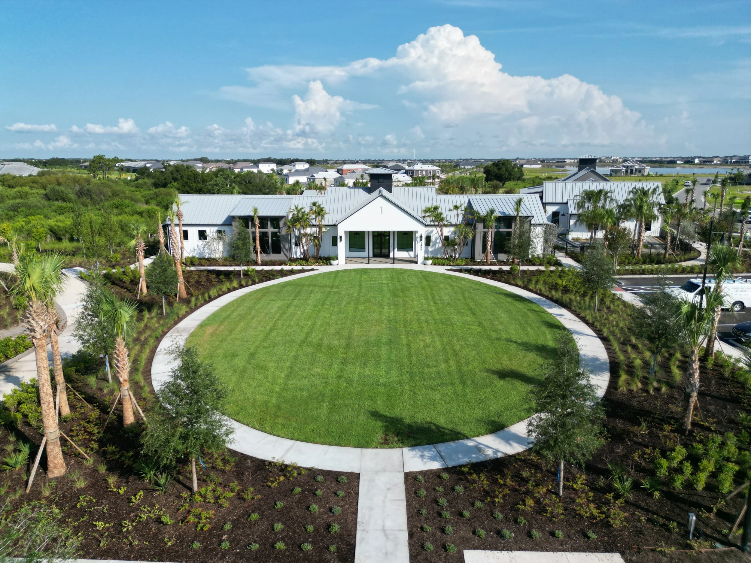 An aerial view of a landscaped residential community with white modern houses, a green lawn, pathways, palm trees, and a partly cloudy sky.