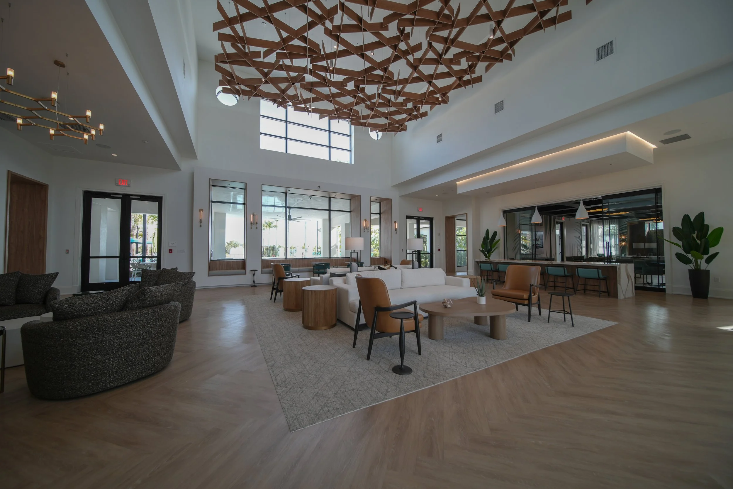 A spacious modern lobby with a high ceiling features a large decorative wooden architectural element hanging from the ceiling. The area is furnished with a variety of chairs and sofas, some wooden and some upholstered, arranged around small tables on