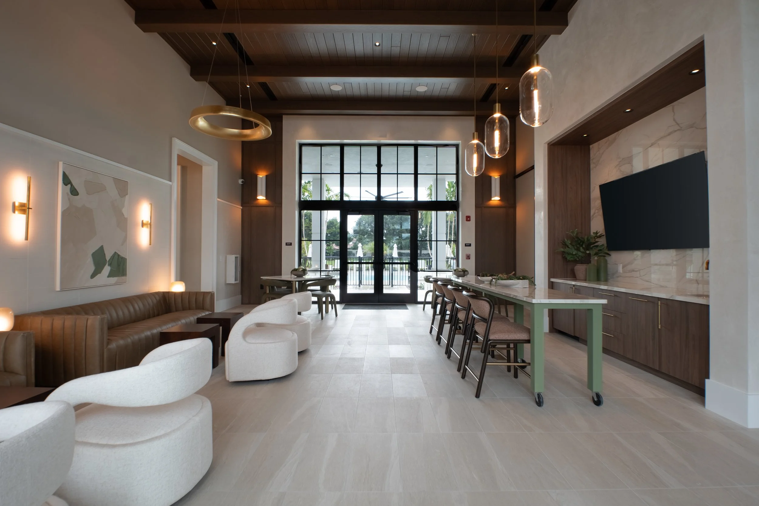 Modern indoor lounge area with beige and white curved chairs, a brown leather sofa, abstract wall art, pendant lighting, large glass windows, and a kitchen island with bar stools.