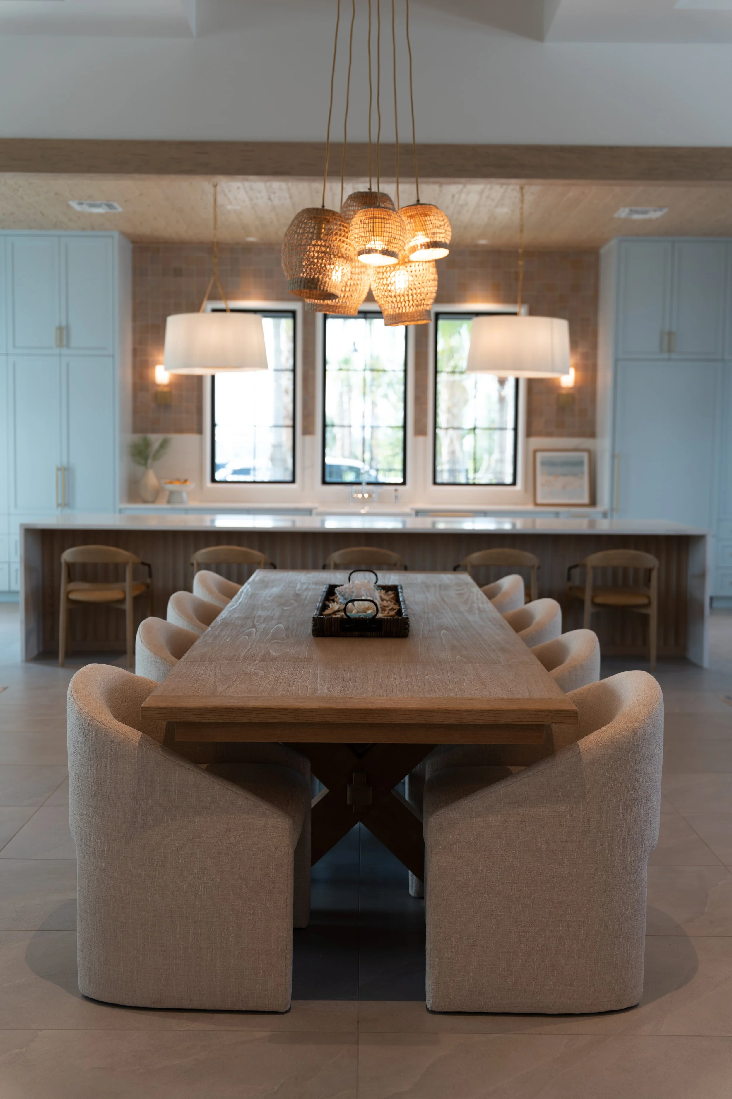 A modern dining room with a large wooden table surrounded by beige upholstered chairs. Above the table hangs a cluster of woven pendant lights. There are two large windows in the background with white frames, flanked by light blue cabinets, and a bar
