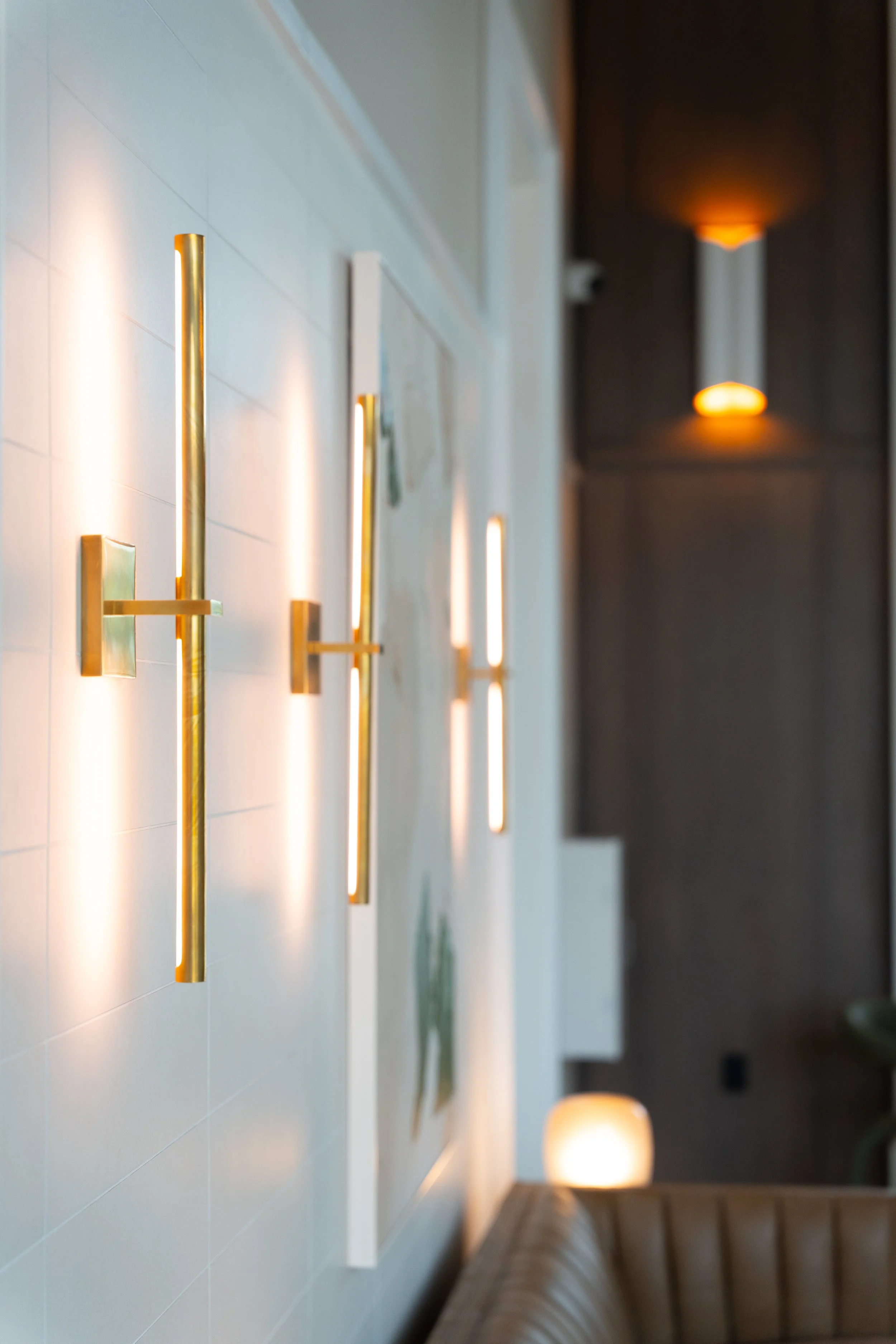 Modern interior hallway with vertical golden wall sconces emitting warm light, white tiled flooring, dark wall with glowing orange lights, and a brown leather bench along the right side.