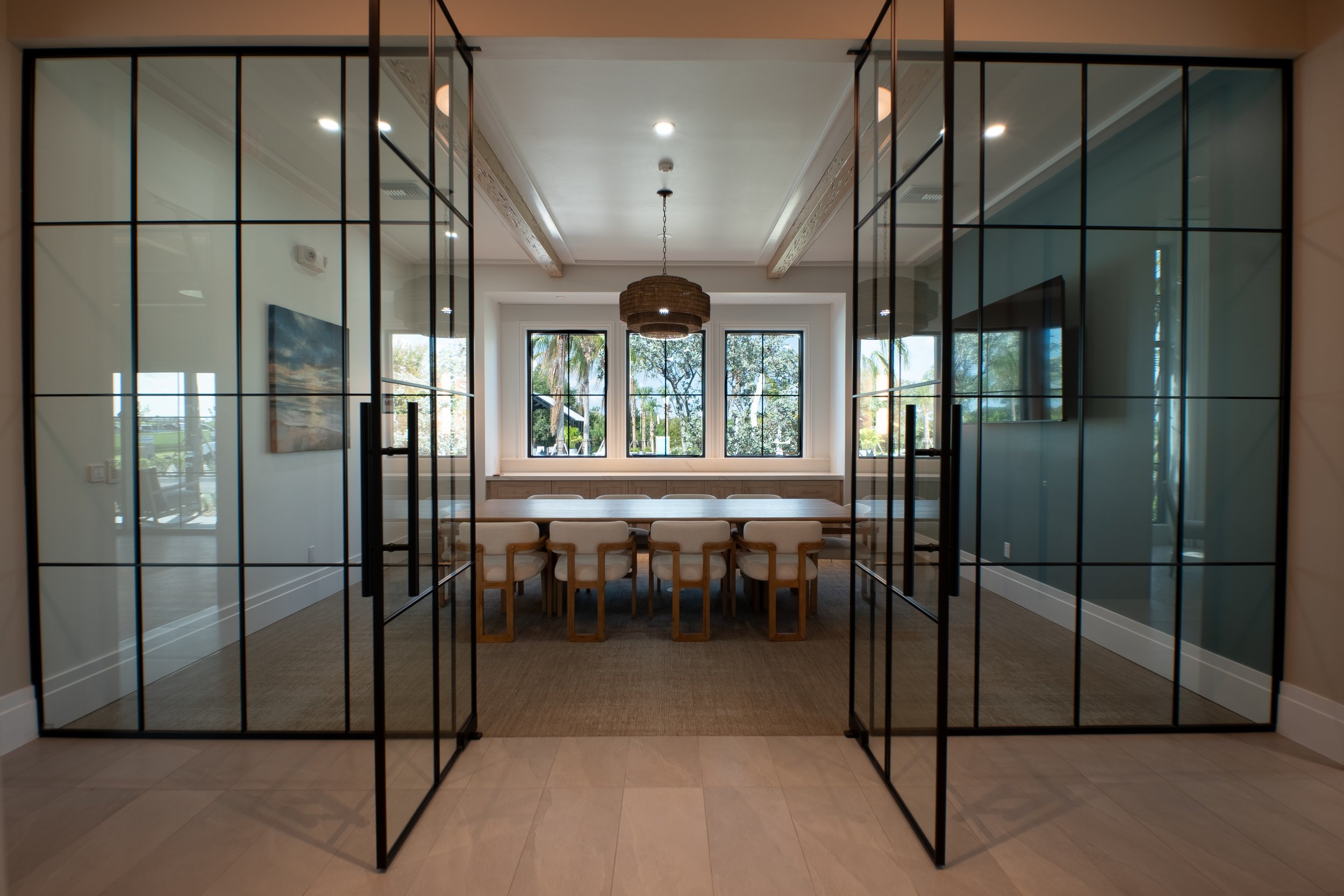 View through black-framed glass sliding doors into a bright dining room with a wooden table and beige chairs, large windows showing trees outdoors, and a woven pendant light hanging from the ceiling.