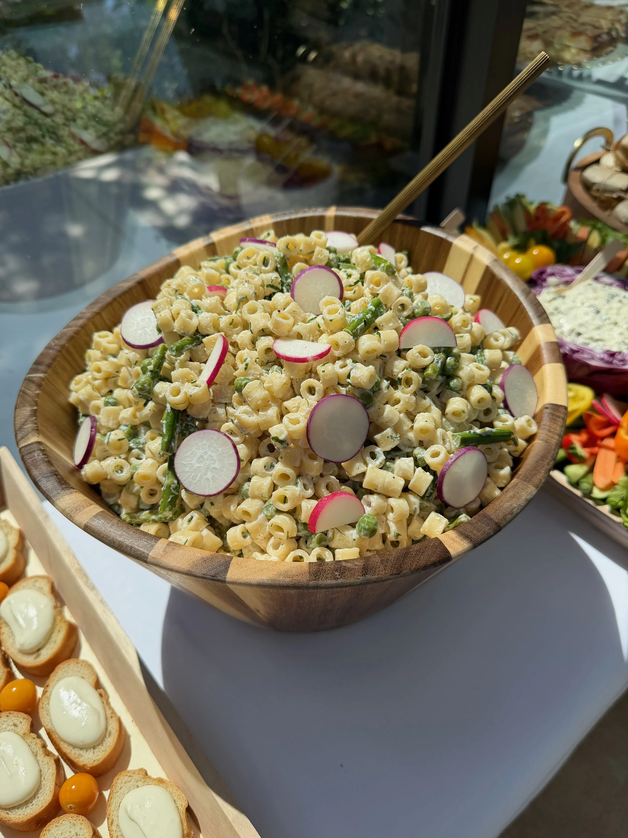 A wooden bowl filled with macaroni salad garnished with sliced radishes and green beans, placed on a white surface. Other dishes with appetizers are partially visible nearby.