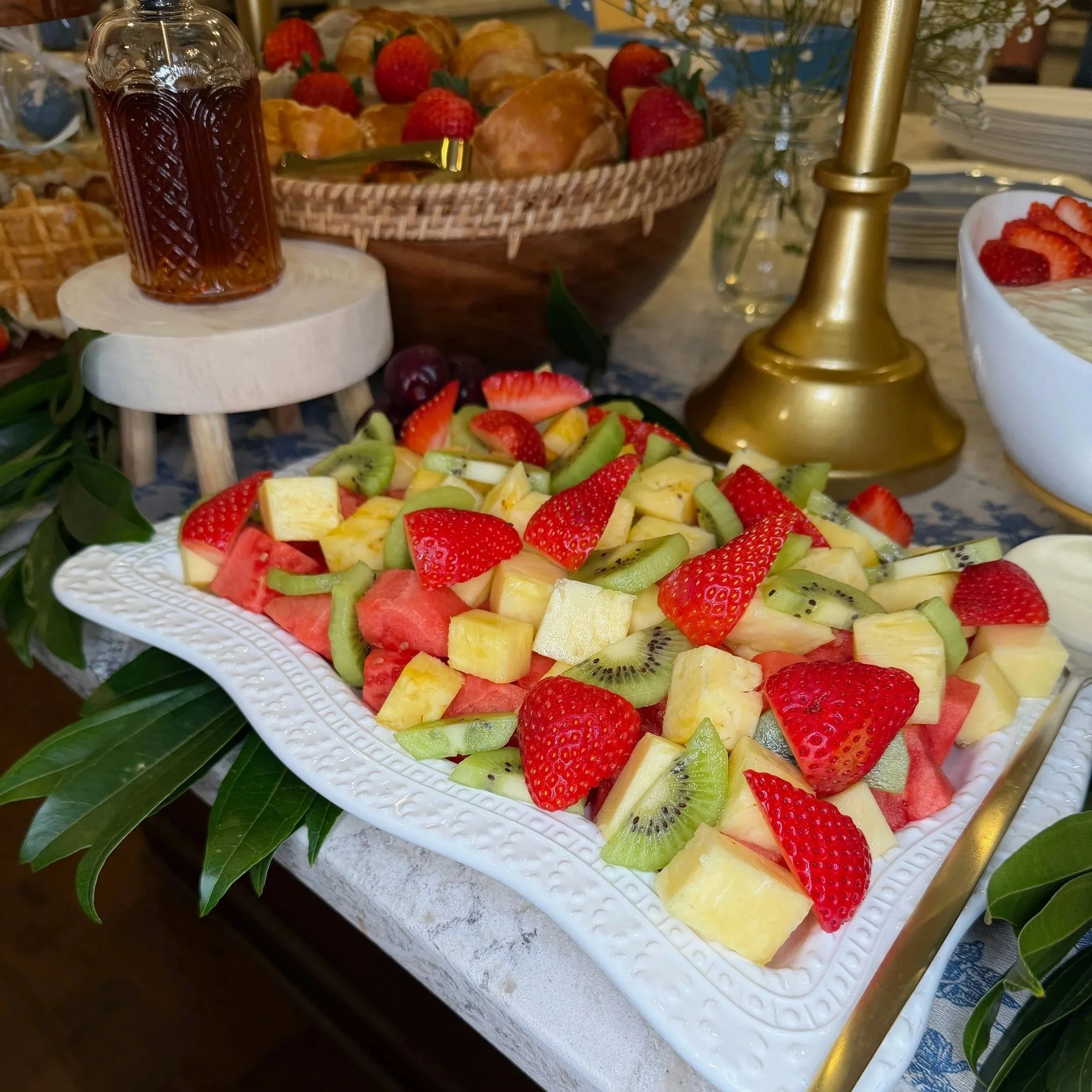 A fruit platter with strawberries, kiwi, pineapple, and watermelon, alongside a jar of syrup on a decorative white tray.