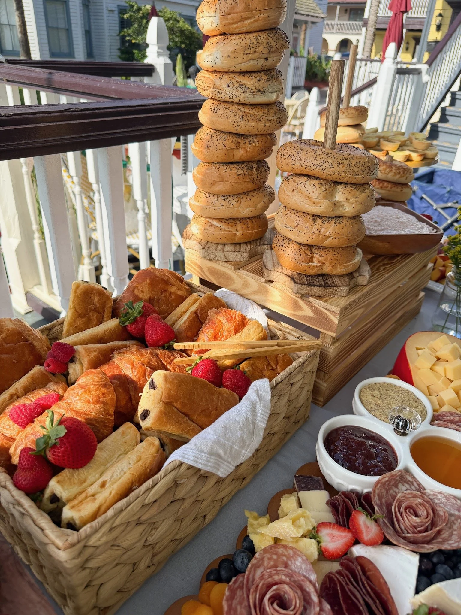 A display of breakfast foods including bagels, pastries, strawberries, blueberries, cheese, ham, and various jams in small bowls at an outdoor setting.