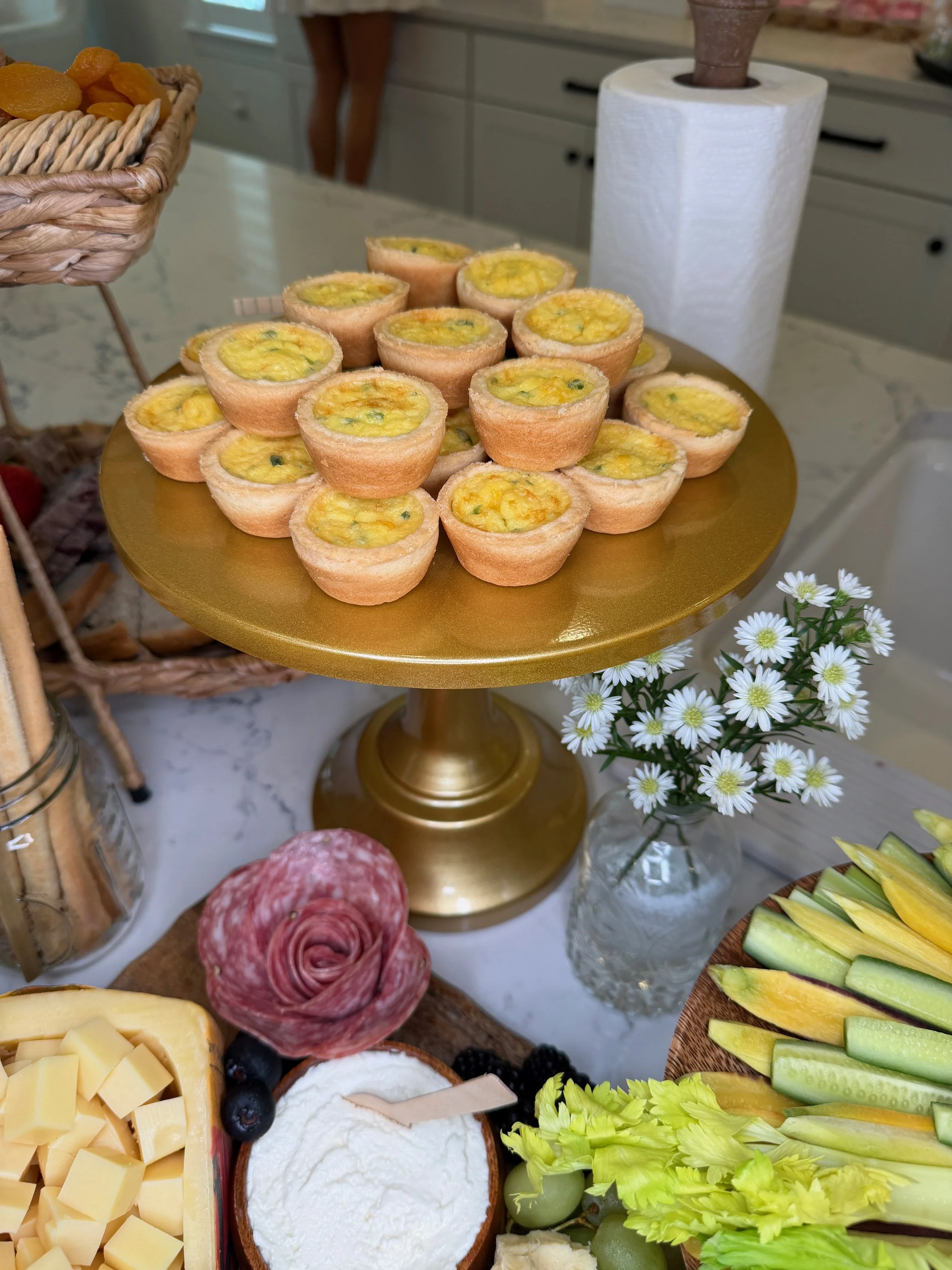 A gold cake stand with bite-sized cheese quiches, a basket of dried apricots, a jar of breadsticks, a cheese platter with cubed cheese, sliced cheese, and salami, a bowl of strawberries and blueberries, a bowl of whipped cream, fresh vegetables inclu