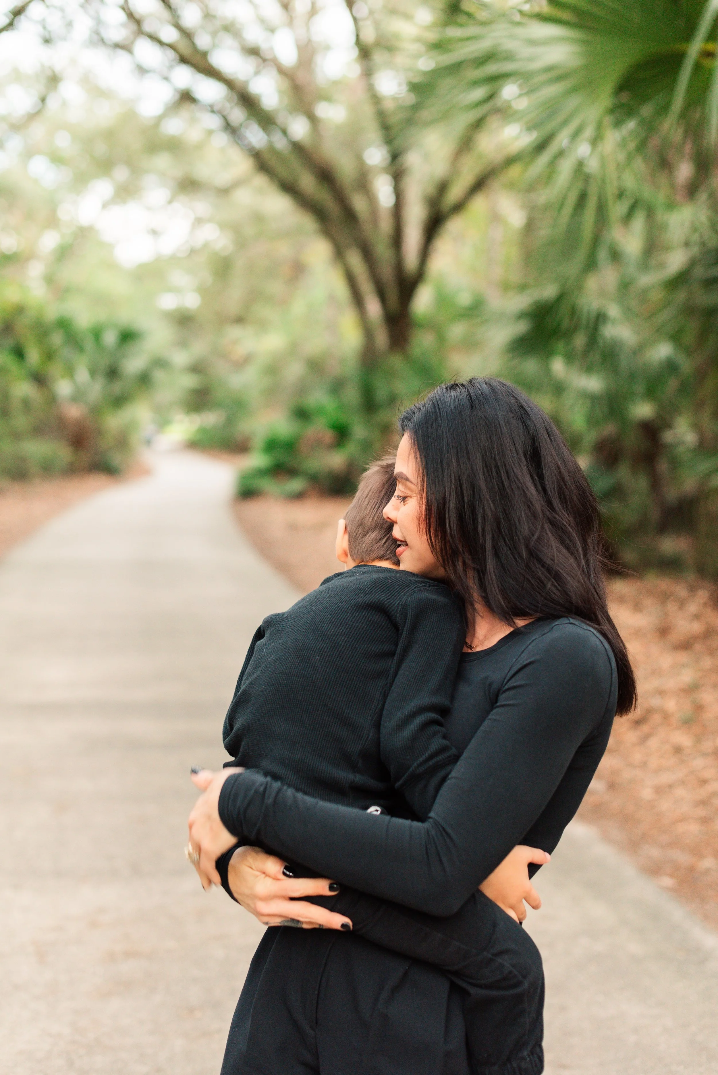 A woman with black hair hugging a young boy outdoors on a pathway surrounded by green trees.