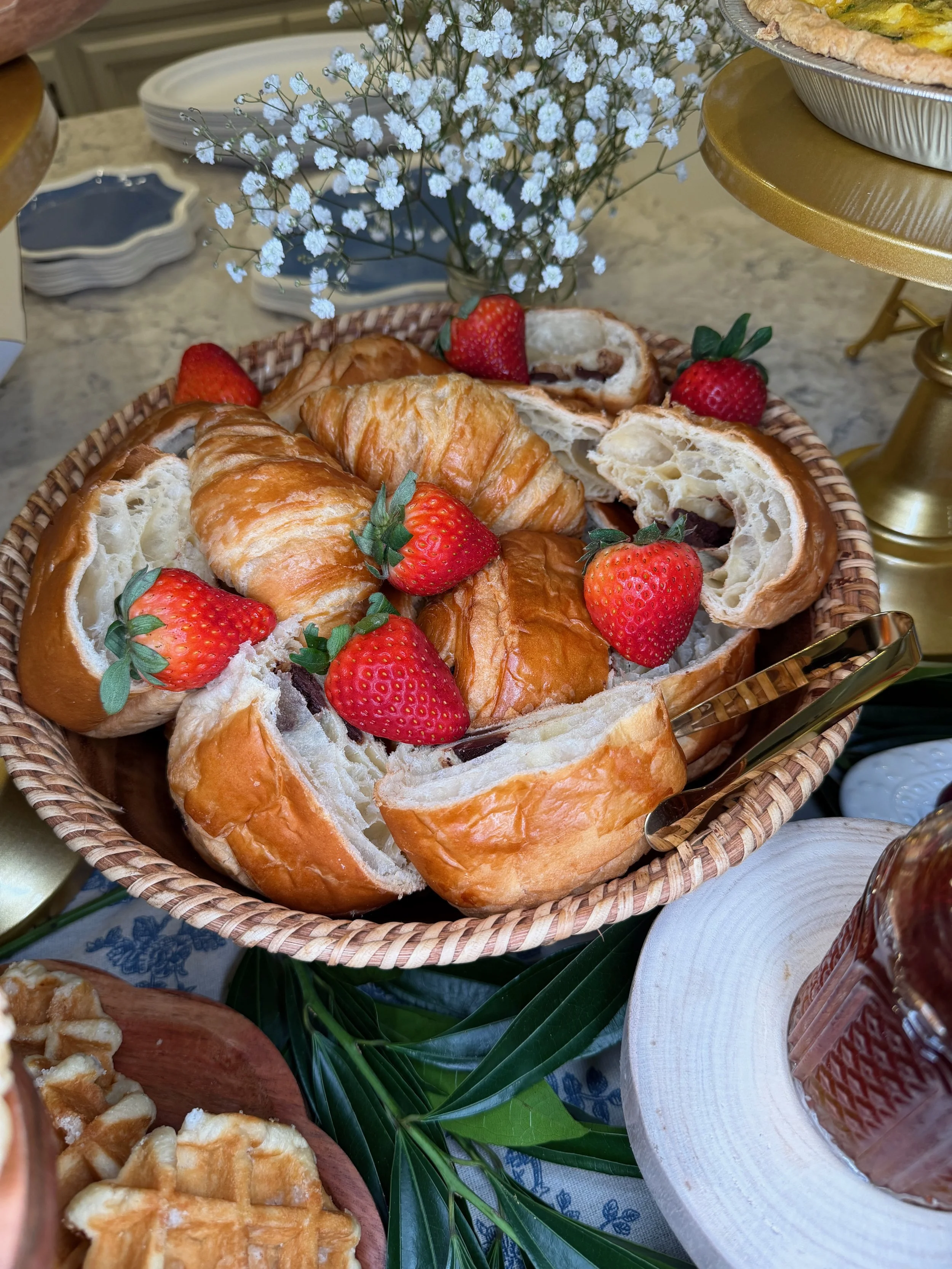 A basket of croissants with strawberries on top, part of a dessert spread on a table with flowers and plates.