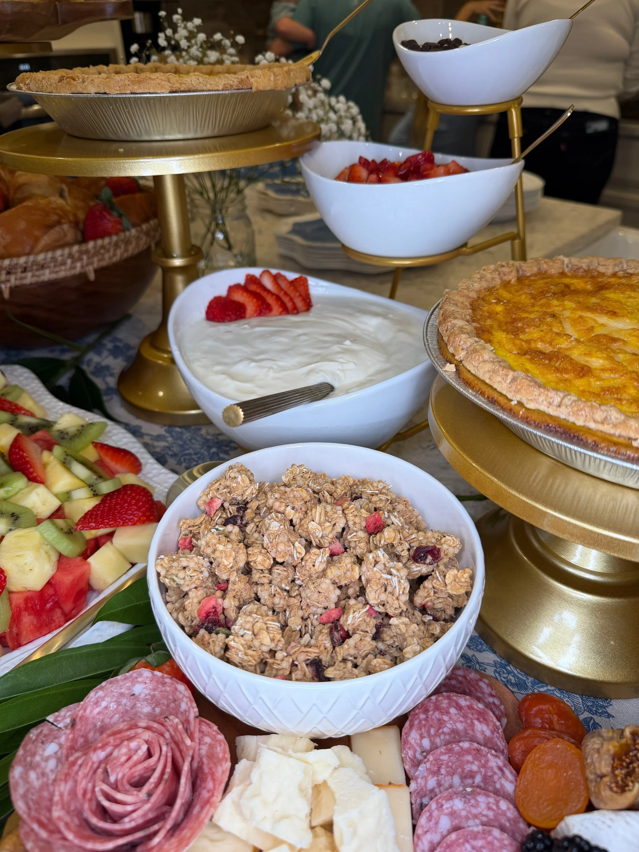 A buffet table with various food items including sliced cured meats, cheeses, fresh fruits, granola, yogurt, and desserts, arranged on gold stands and white bowls.