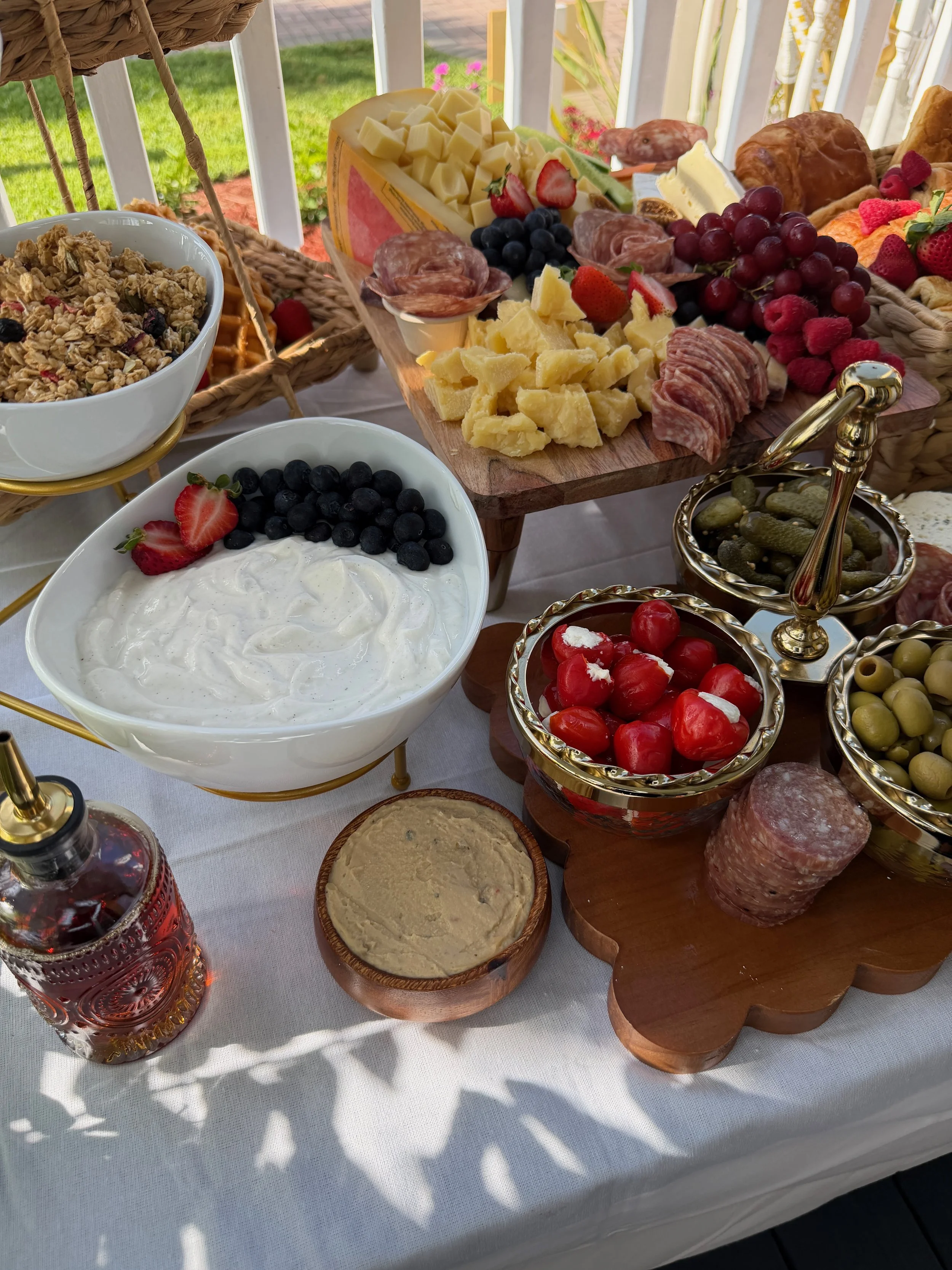 A breakfast or brunch spread on a table outdoors with various cheeses, meats, fresh berries, granola, and pickles.