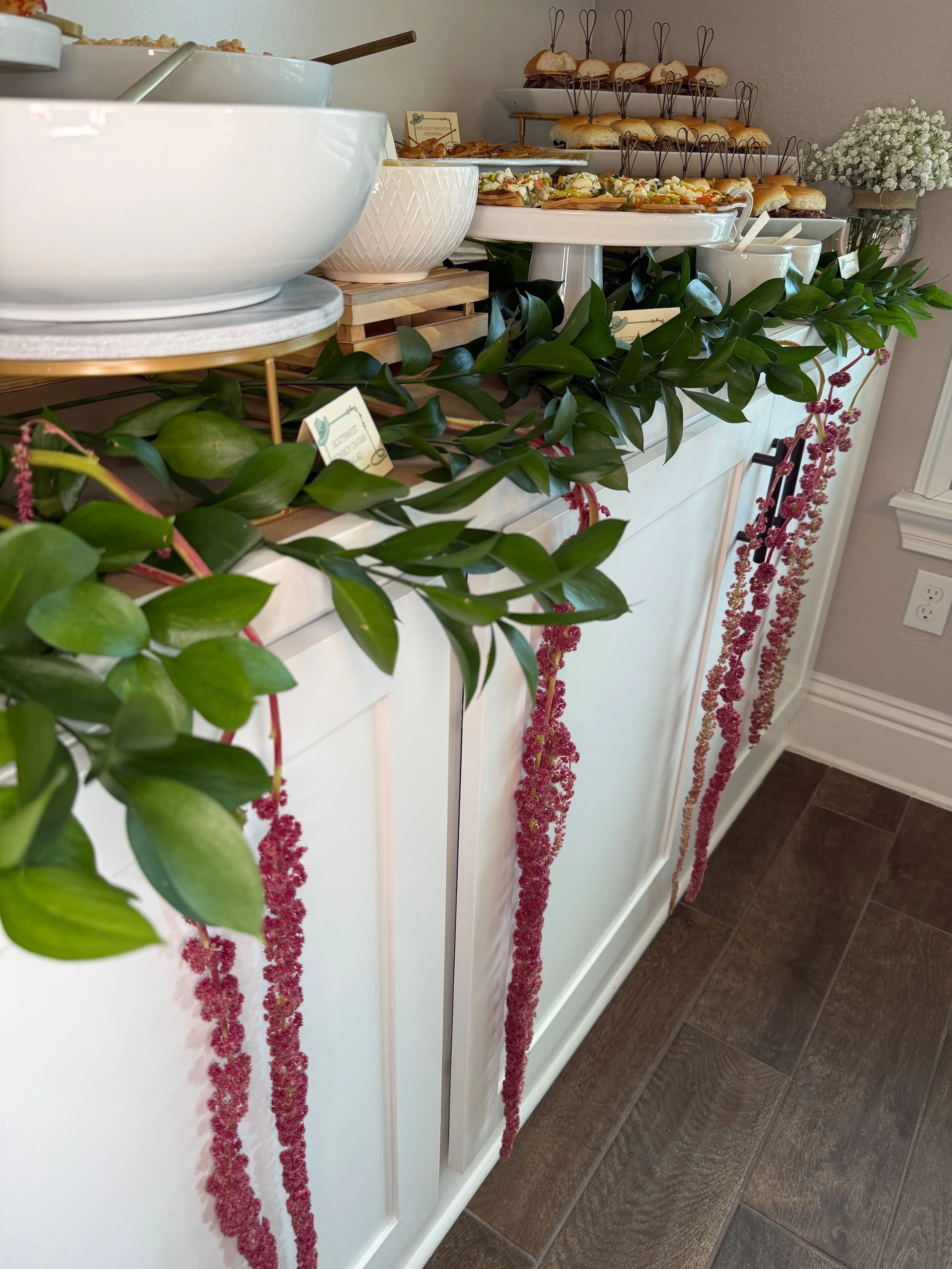 Display of assorted sandwiches, pastries, and salads on tiered platters, decorated with greenery and floral garlands, in a buffet setting.