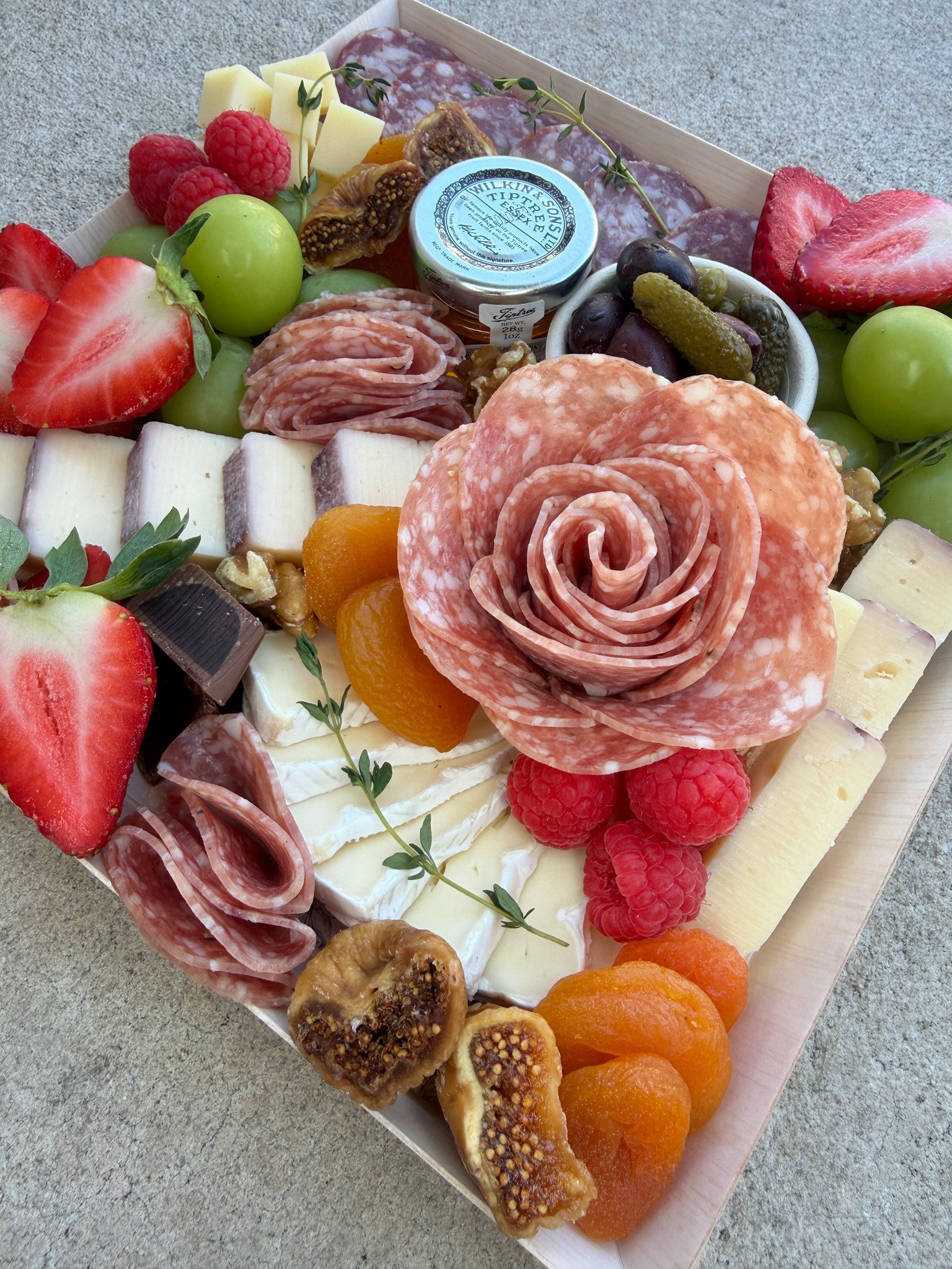 A tray of assorted cheeses, cured meats, fresh fruit including strawberries, grapes, raspberries, figs, dried apricots, and a small jar of preserves.