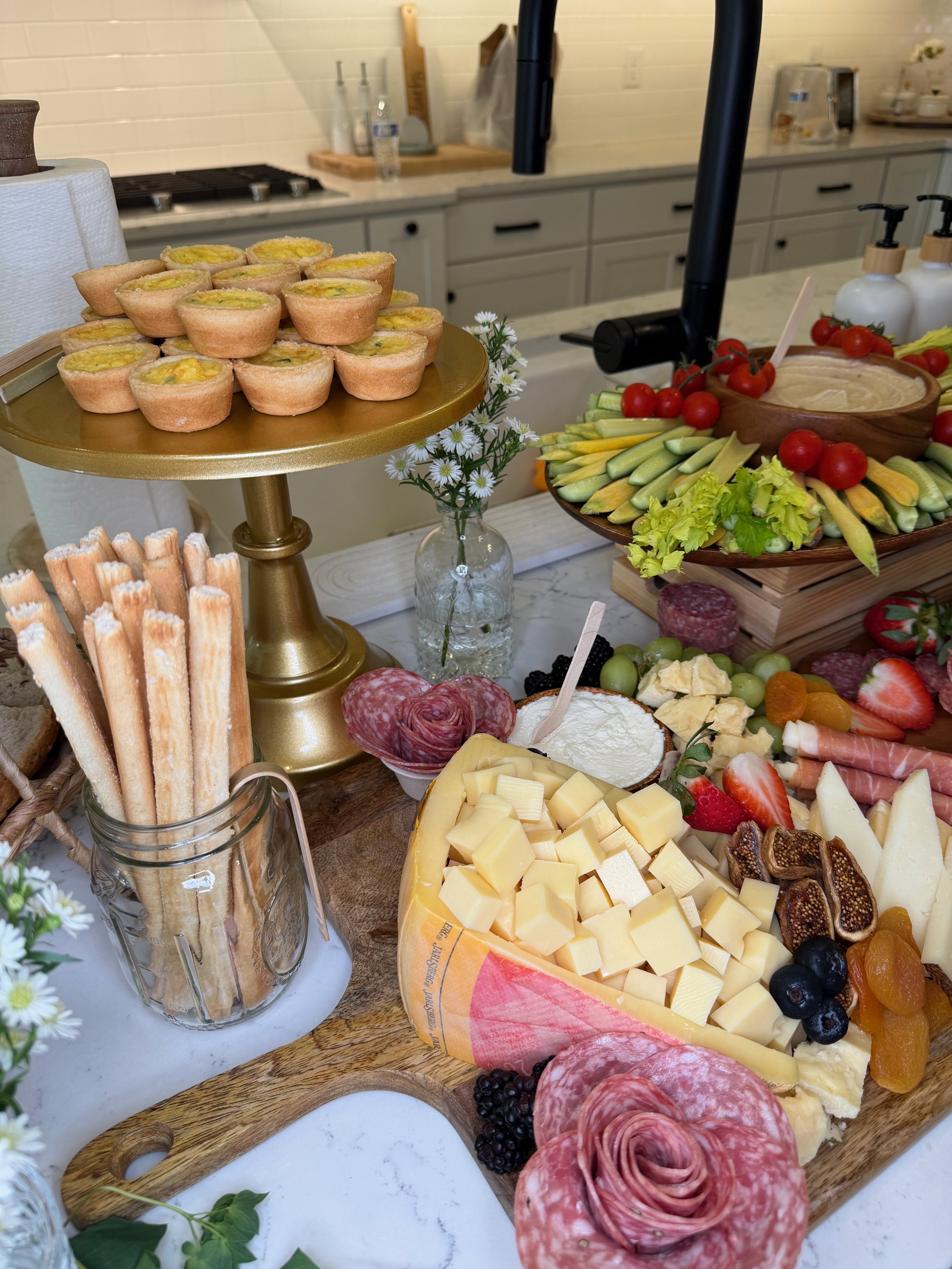 Assorted cheeses, sliced meats, fresh vegetables, cherry tomatoes, and crackers on a charcuterie board and serving platters at a buffet table.