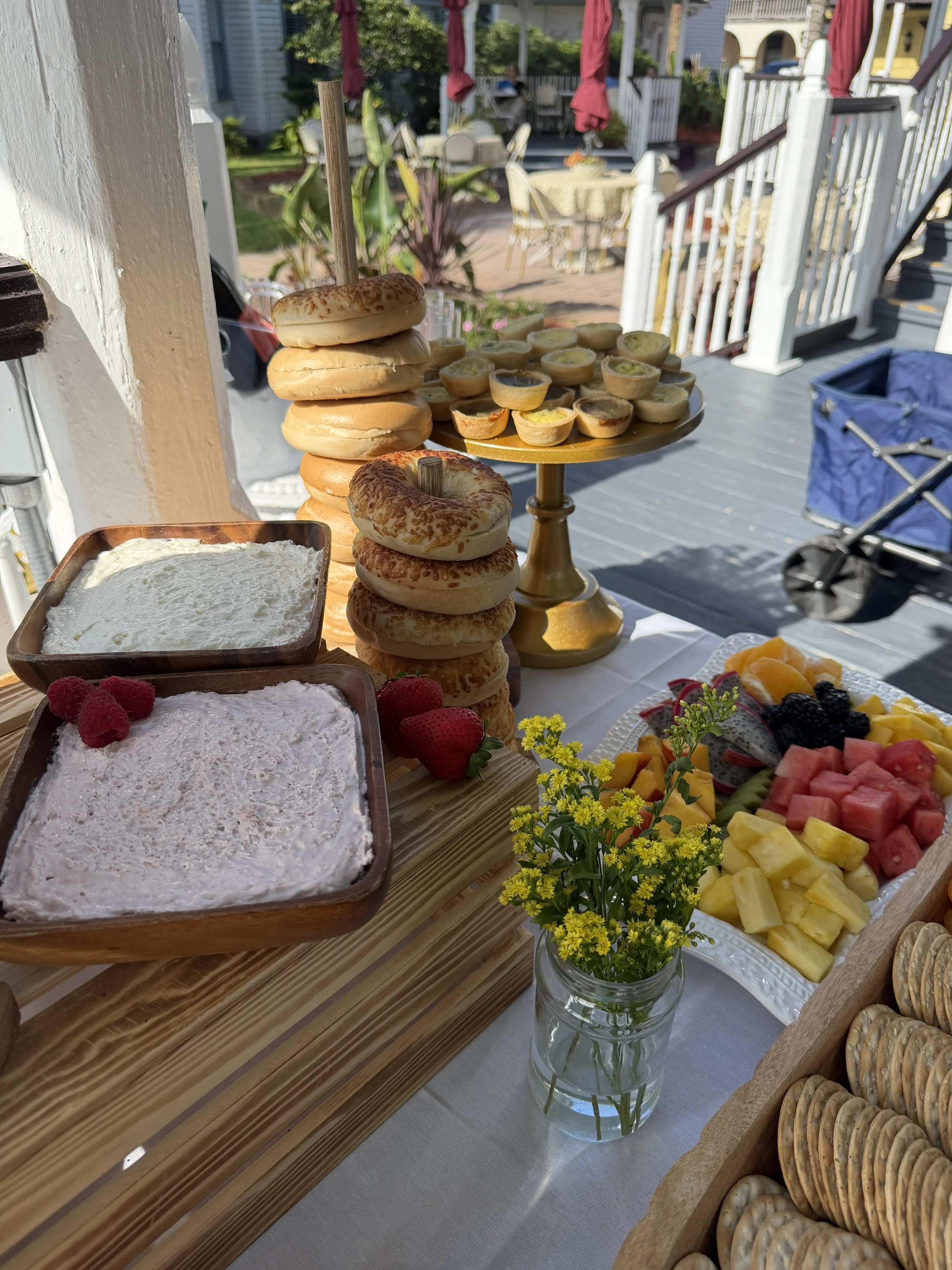 A buffet table set with mini quiches, bagels with cream cheese, and fresh fruit including strawberries, blackberries, pineapple, watermelon, and cantaloupe. There is a vase with yellow flowers in the foreground, and a porch with outdoor furniture in 