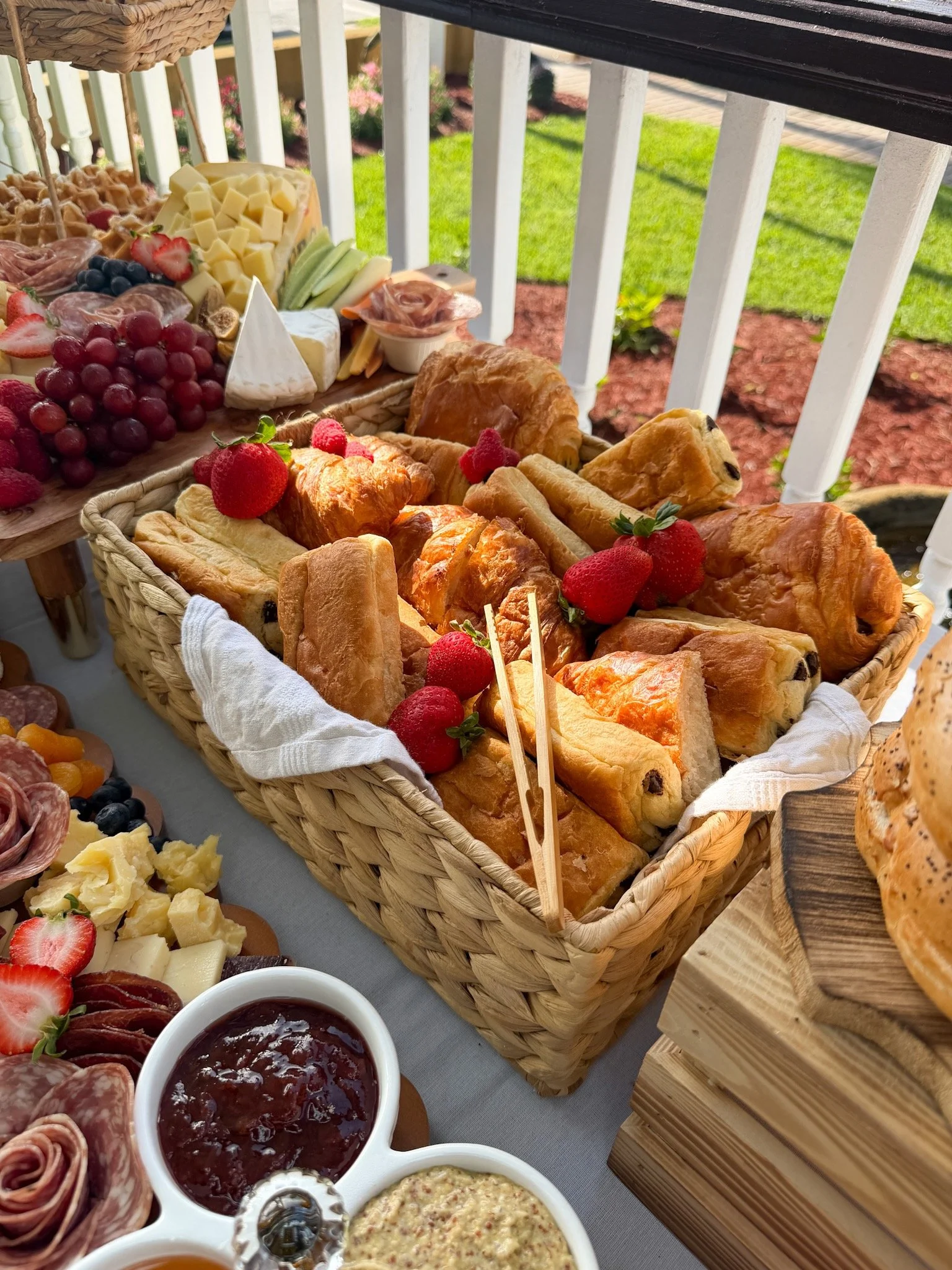 A table set with assortments of baked goods, strawberries, grapes, cheese, charcuterie, and jars of jam or preserves, with a grassy outdoor background.