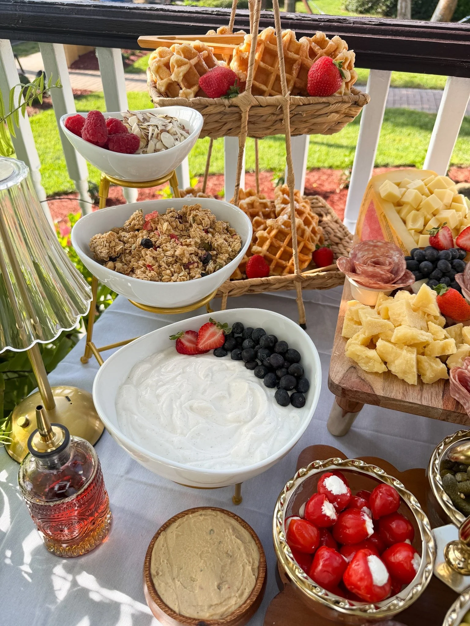 Assorted breakfast items on a table including bowls of yogurt with blueberries, strawberries, and whipped cream, granola, cheese, a basket of waffles with strawberries, and bottles of drink, set outdoors with grass and a white fence in the background