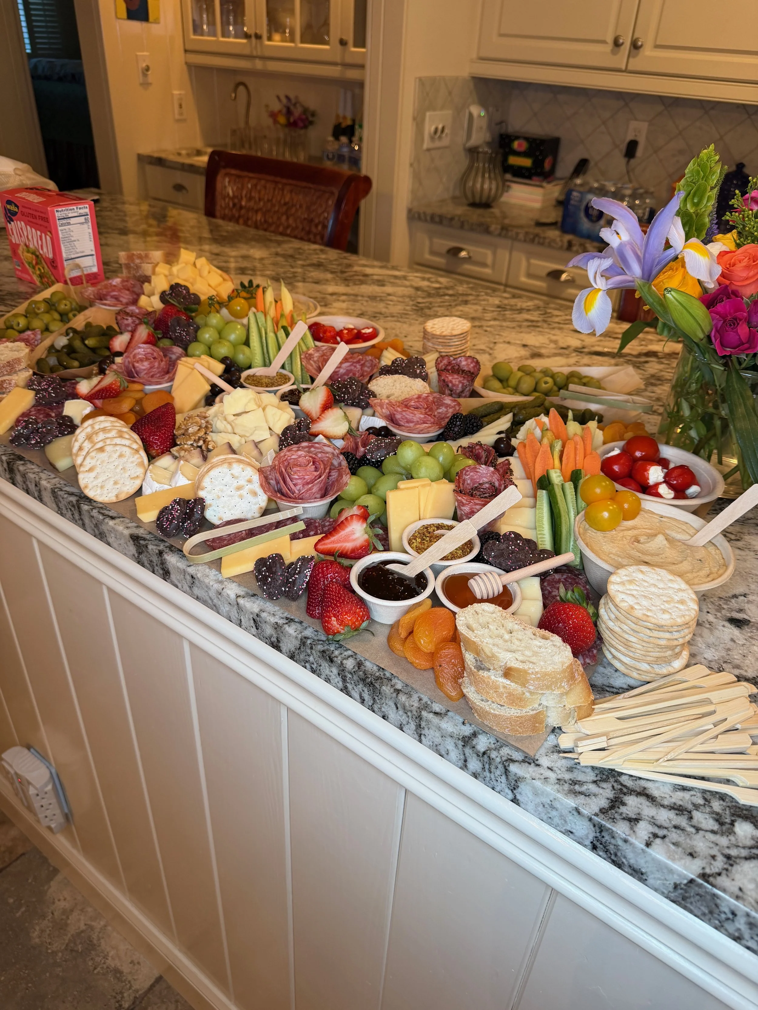 A kitchen island with an extravagant spread of cheese, fruits, crackers, vegetables, and various appetizers, and a vase with flowers.