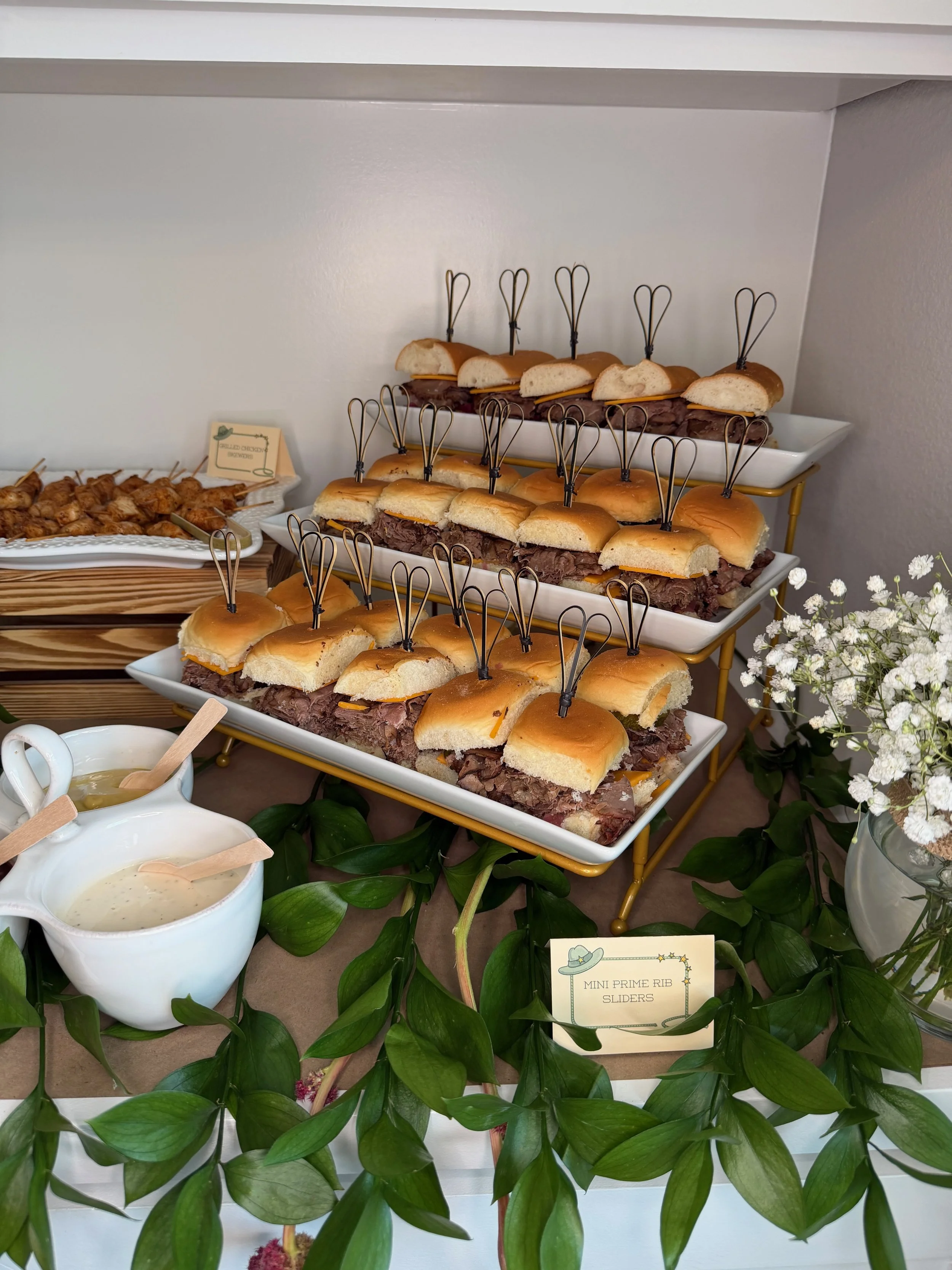 A buffet display featuring mini prime rib sliders on white plates with small black handles, arranged on a tiered golden stand. The sliders have beef and cheese in small sandwich buns. To the left, there are skewers of chicken and a small card that re