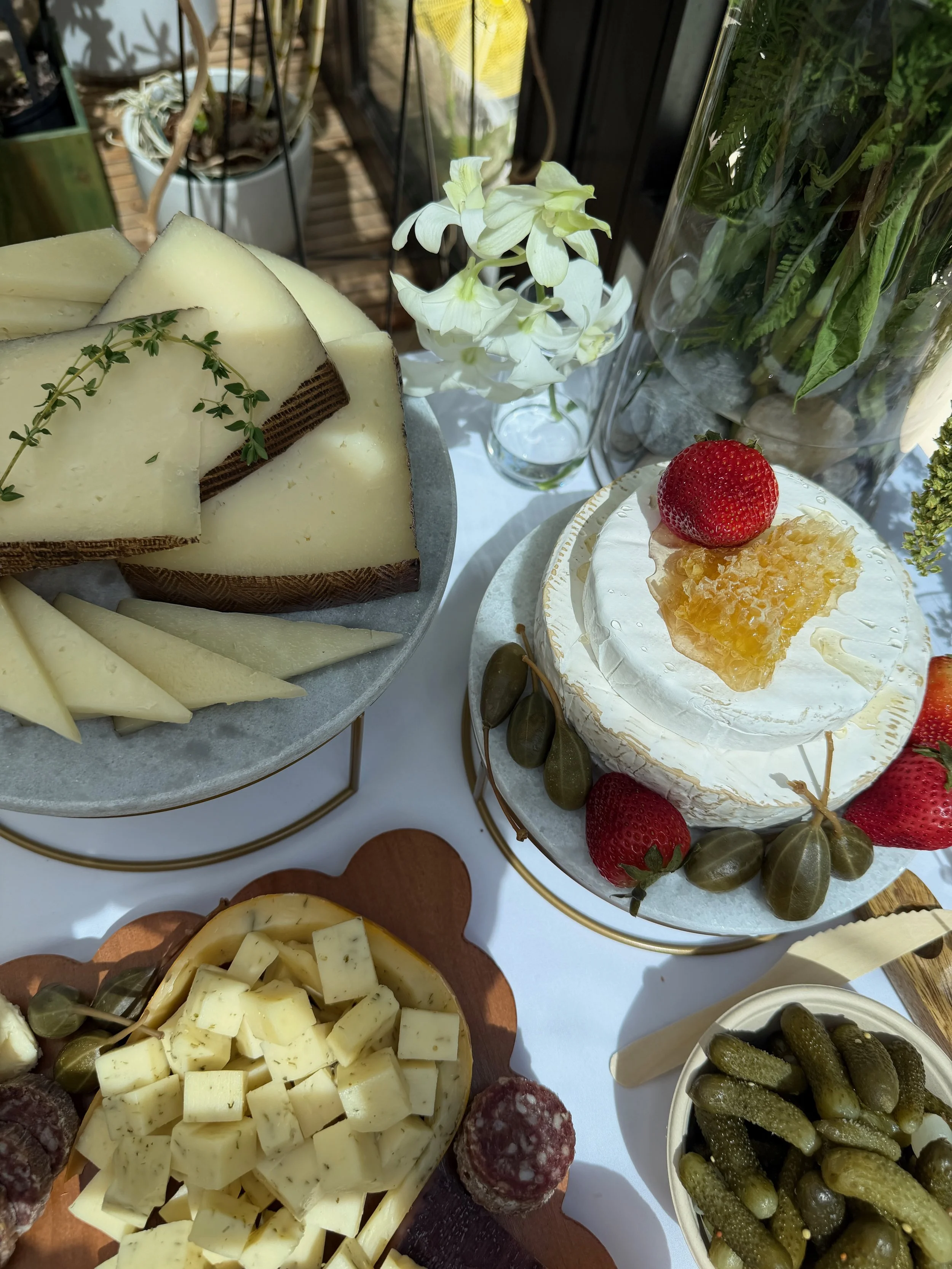 A cheese platter with sliced cheese, herbs, salami, and pickles, alongside a white cake garnished with strawberries, honeycomb, and herbs. The table features decorative elements and flowers.