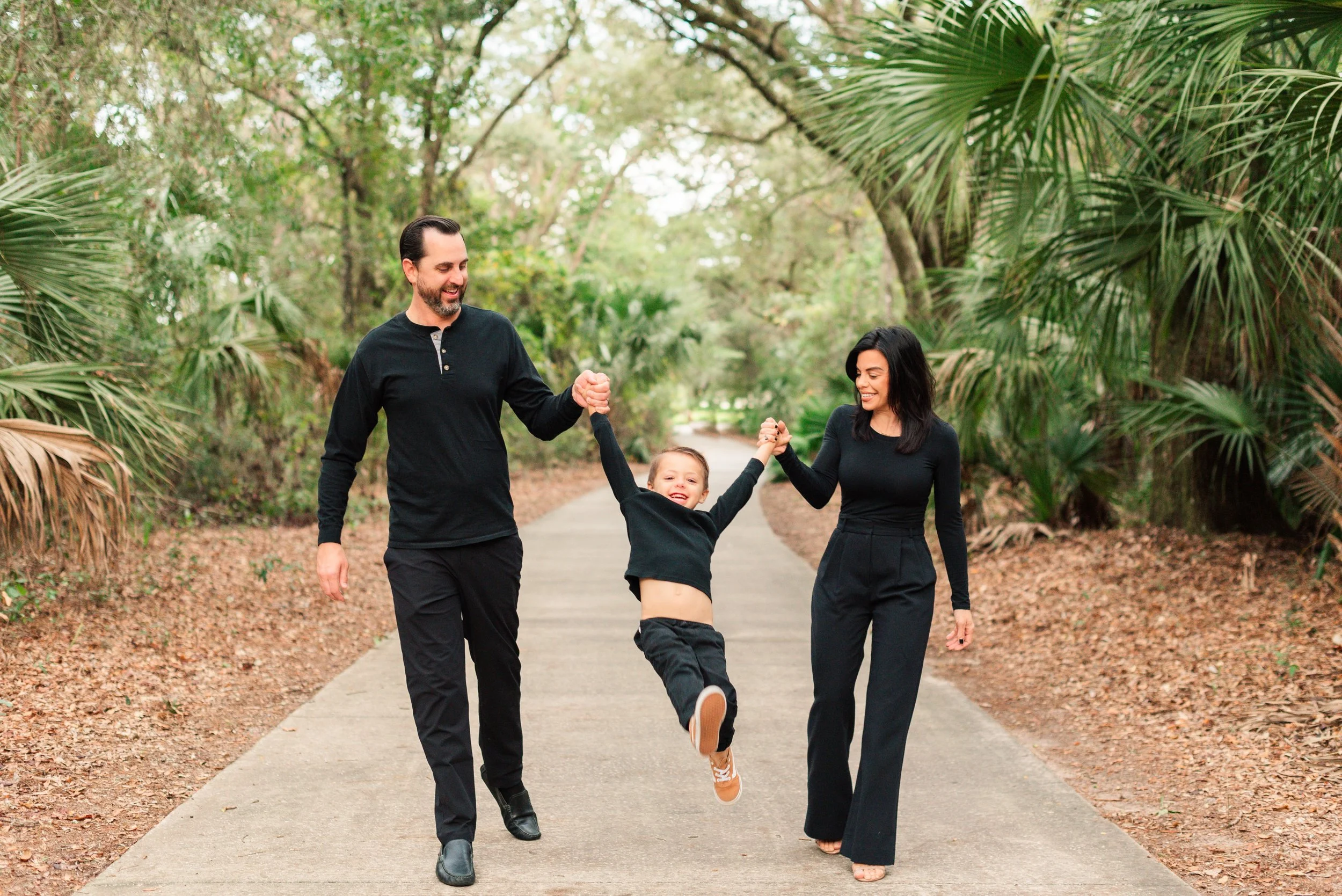 A family of three, two adults and a child, walking on a paved path in a lush, green park. The adults are holding the child's hands, and everyone appears happy. The background shows dense trees and foliage.