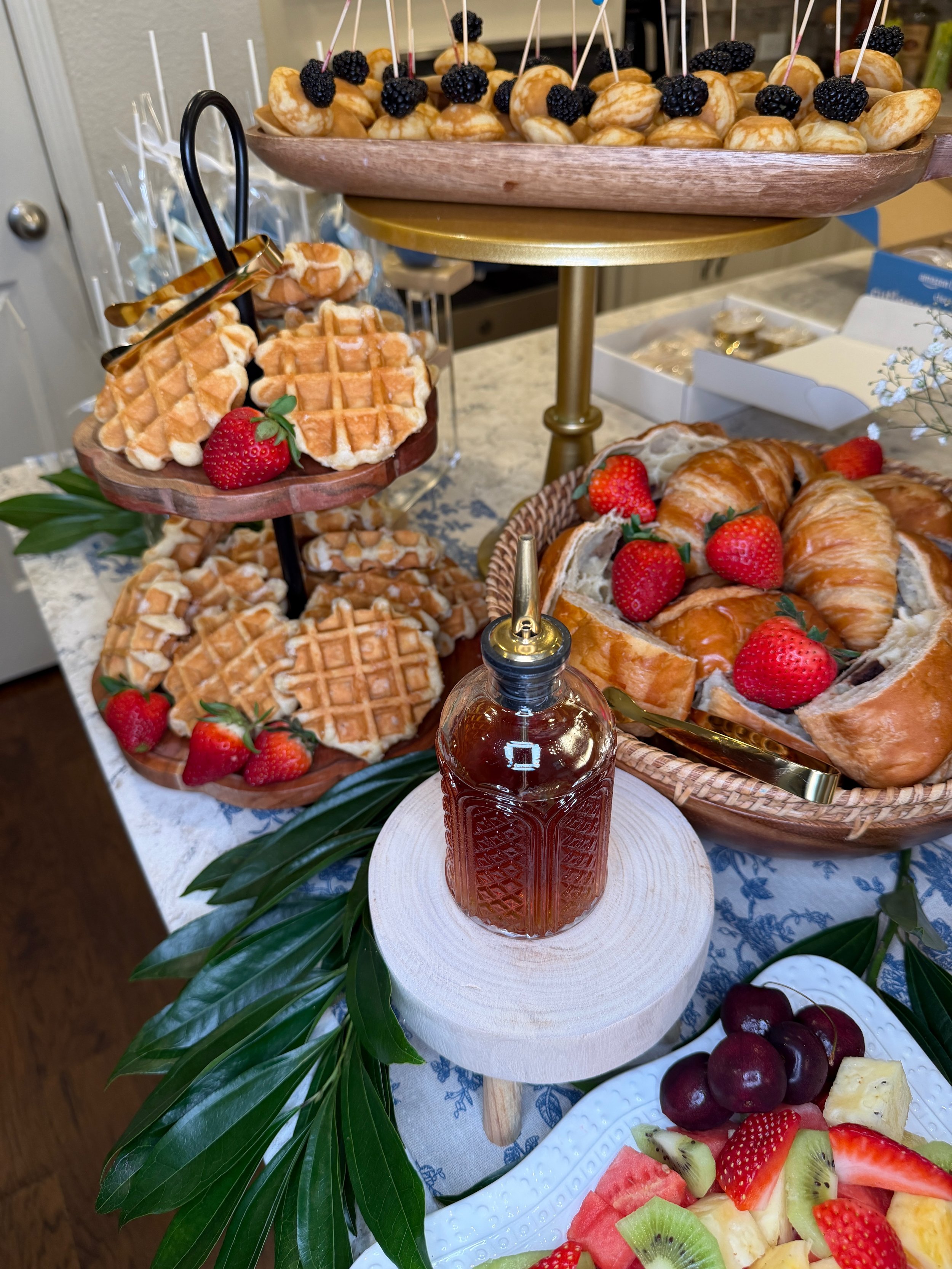 Assorted desserts including waffles with strawberries, mini profiteroles with blackberries, croissants with strawberries, and a fruit platter with watermelon, kiwi, strawberries, and cherries, displayed on a decorative table with a elegant patterned 