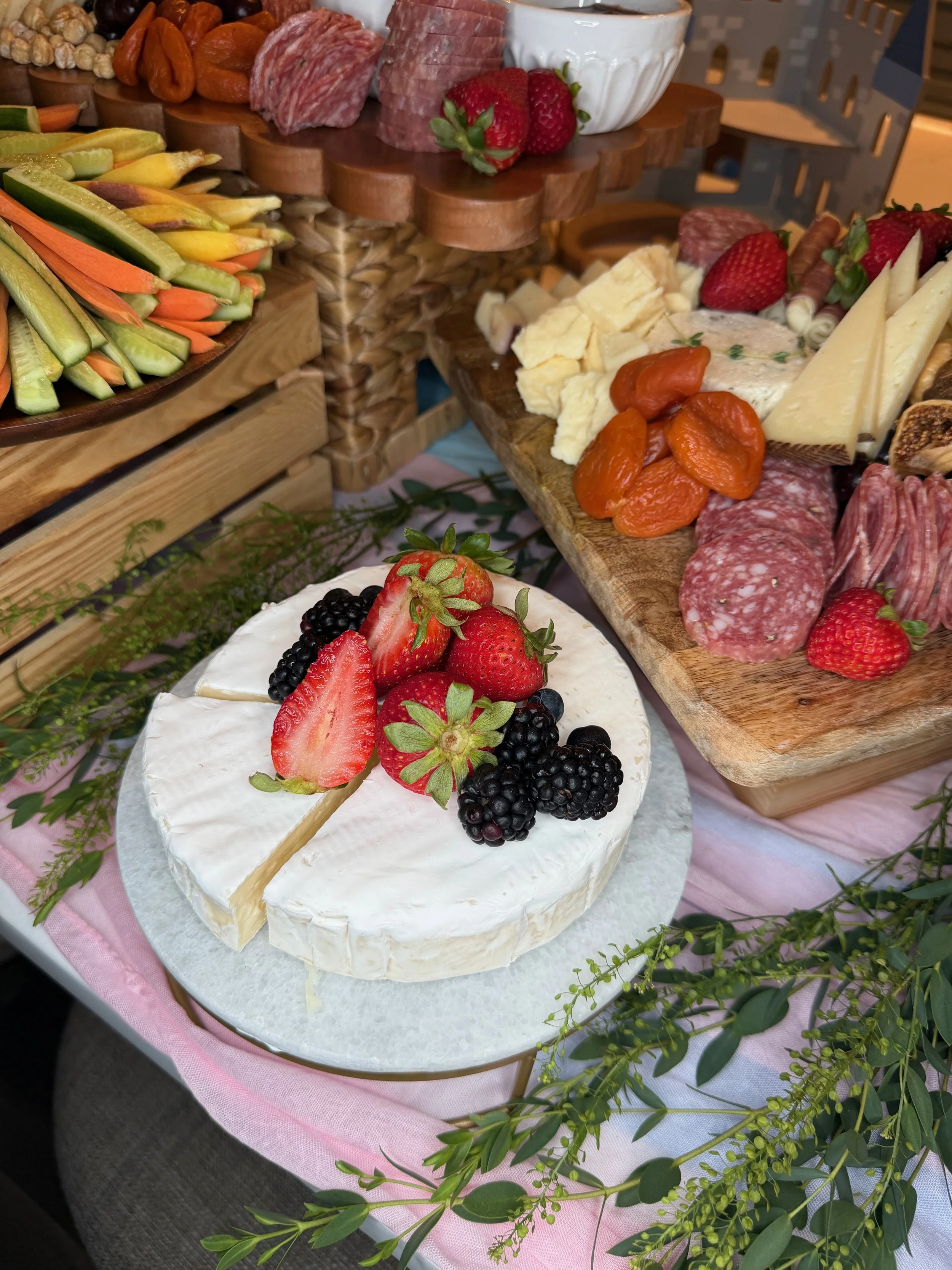 A cheese and meat platter with strawberries, grapes, and blackberries, sitting on a table with greenery and a pink tablecloth.