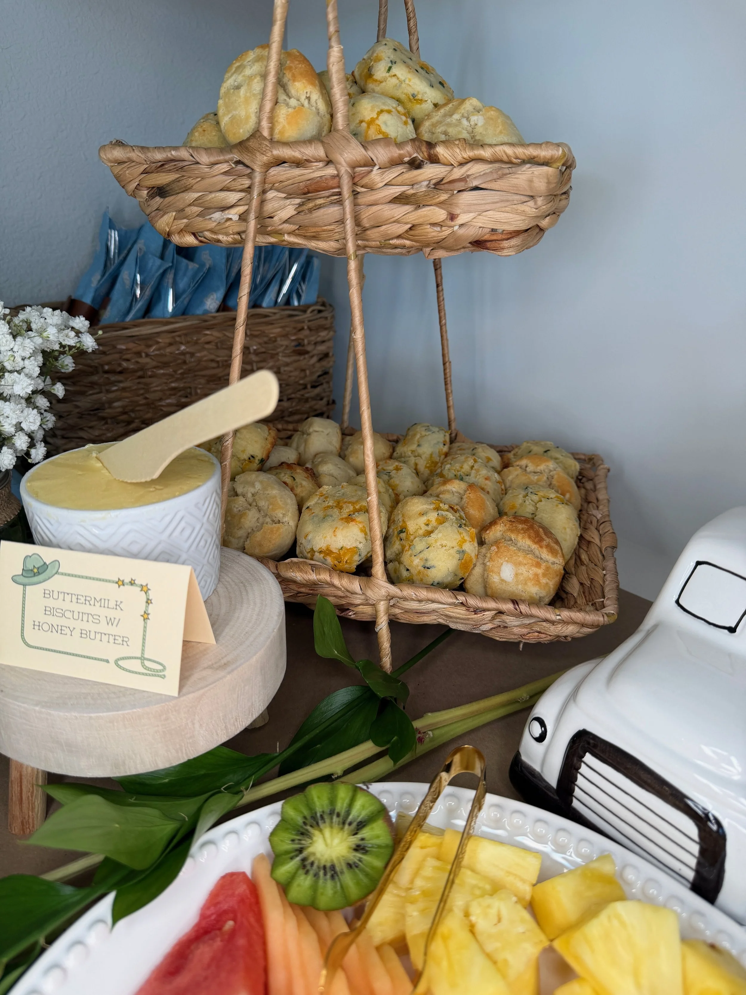 Basket of assorted biscuits on a tiered stand, with a bowl of honey butter and a sign reading 'buttermilk biscuits with honey butter' nearby. Fresh fruit on a white platter in the foreground, including kiwifruit, watermelon, cantaloupe, and pineapple