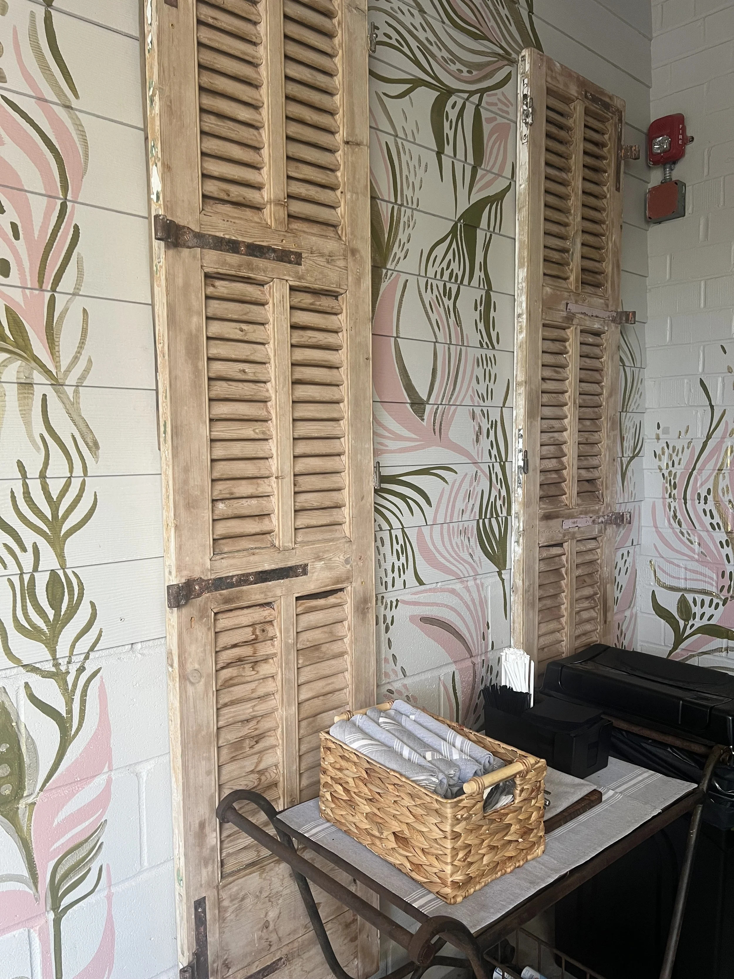 Decorative wall with painted floral and leaf pattern, wooden window shutters, and a small table with a basket, black container, and a citrus squeezer.