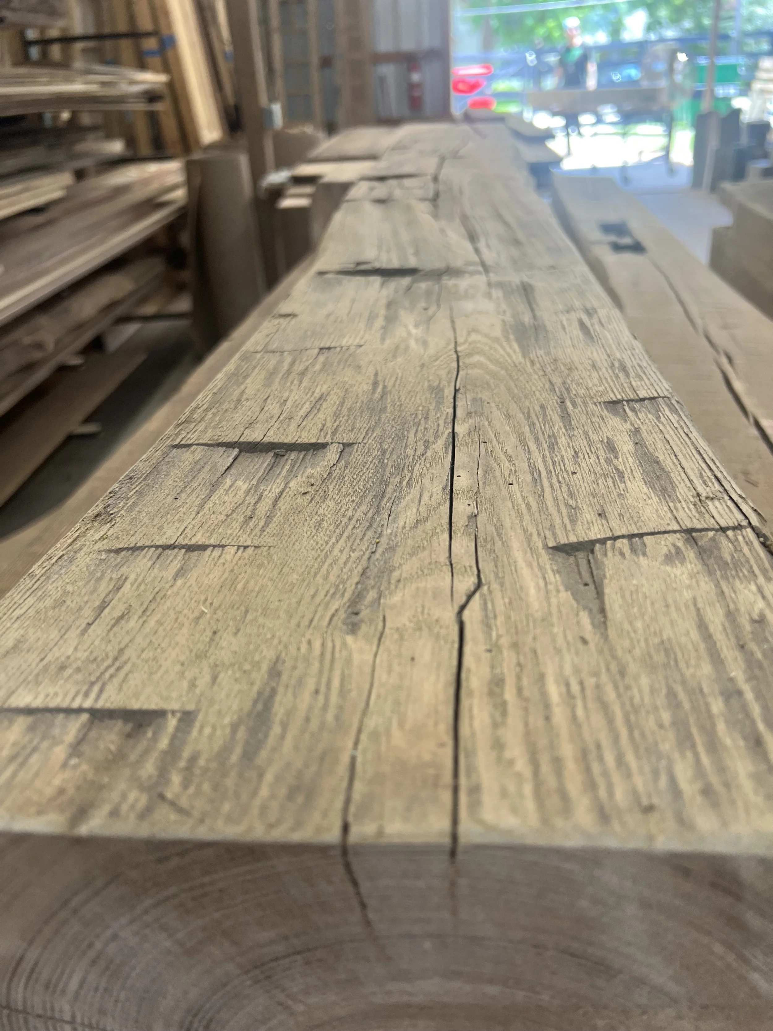 Close-up of a wooden plank with visible grain and cracks, in a woodworking or hardware store with shelves of other wood planks in the background.