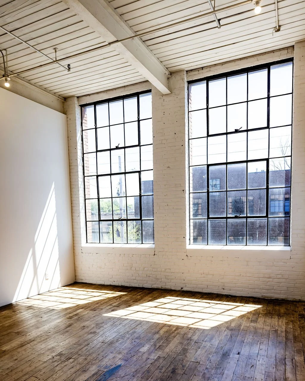 Empty loft with two large industrial-style windows, white brick walls, wooden flooring, and sunlight casting shadows through the windows.