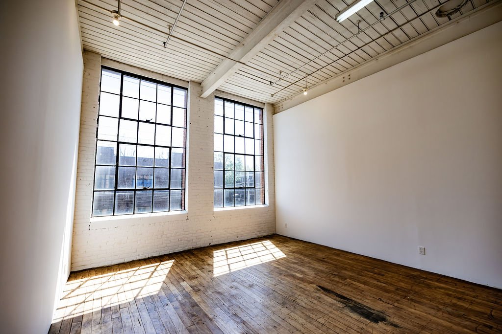 Empty industrial-style room with large windows, white brick wall, wood flooring, and a high ceiling with wooden beams and track lighting.