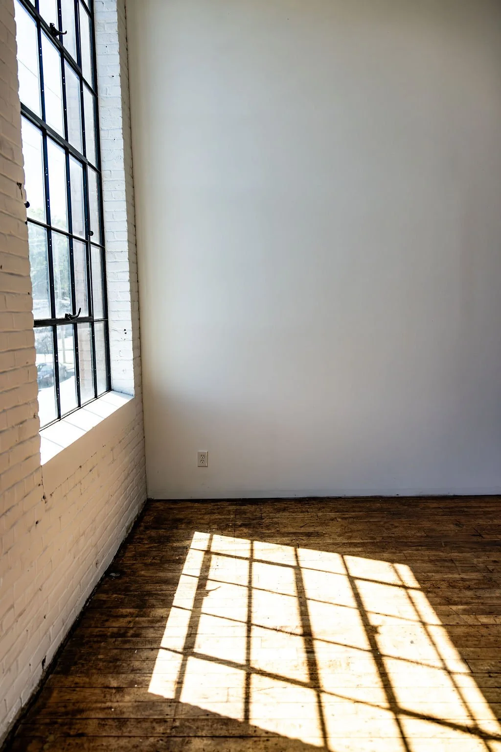 Sunlight streaming through a large industrial-style window, casting a shadow on a wooden floor in a minimalistic, empty room with white walls and an exposed brick wall.