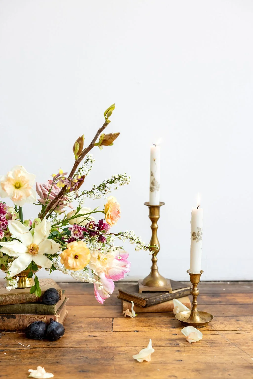 A floral arrangement with various flowers in a vase, placed on a wooden table with some scattered flower petals, and two lit white candles in brass candlesticks.