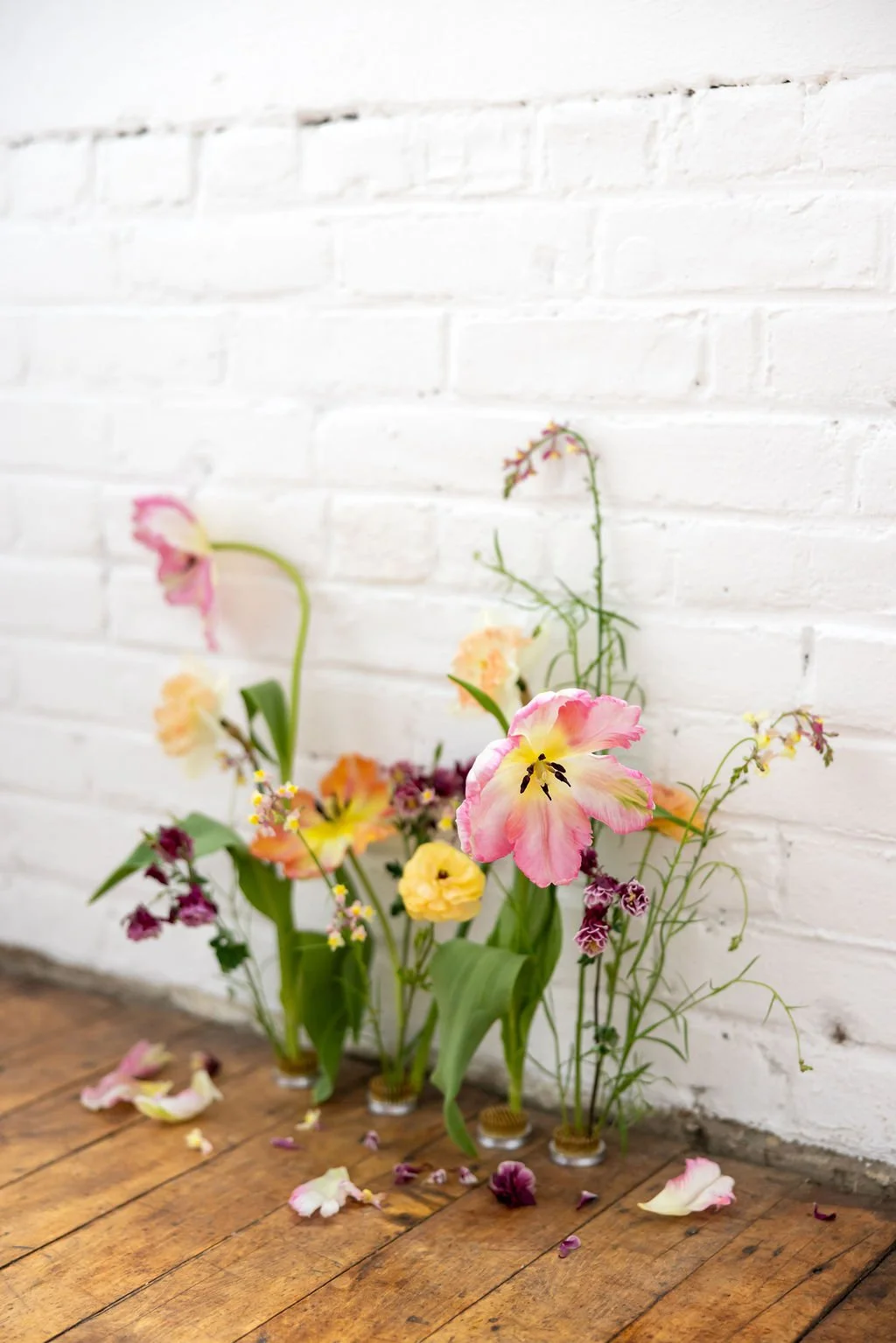 An arrangement of various colorful flowers in small glass vases, placed against a white brick wall and on a wooden floor with scattered flower petals.