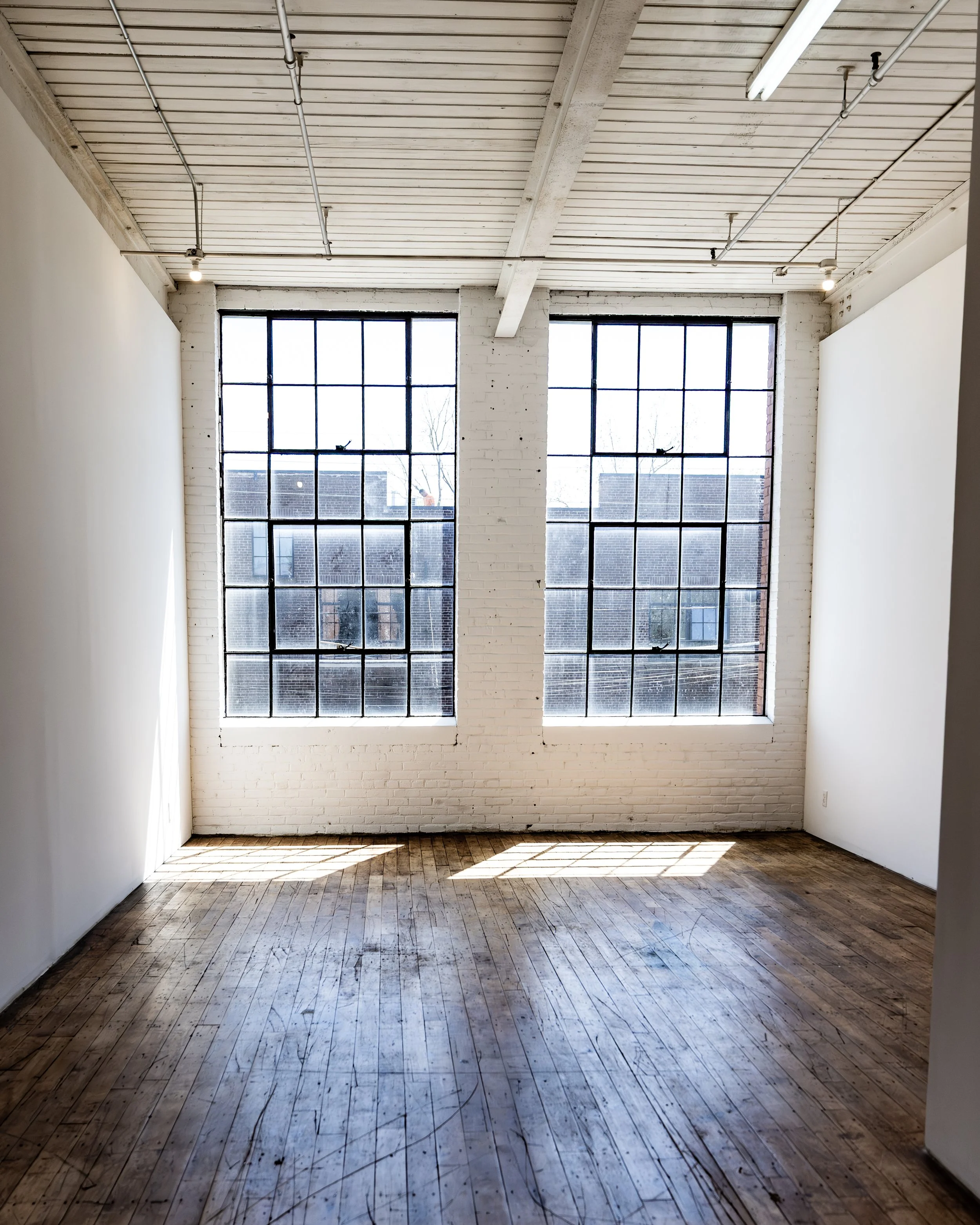 Empty room with large grid windows, white brick walls, wooden floor, and exposed ceiling pipes.