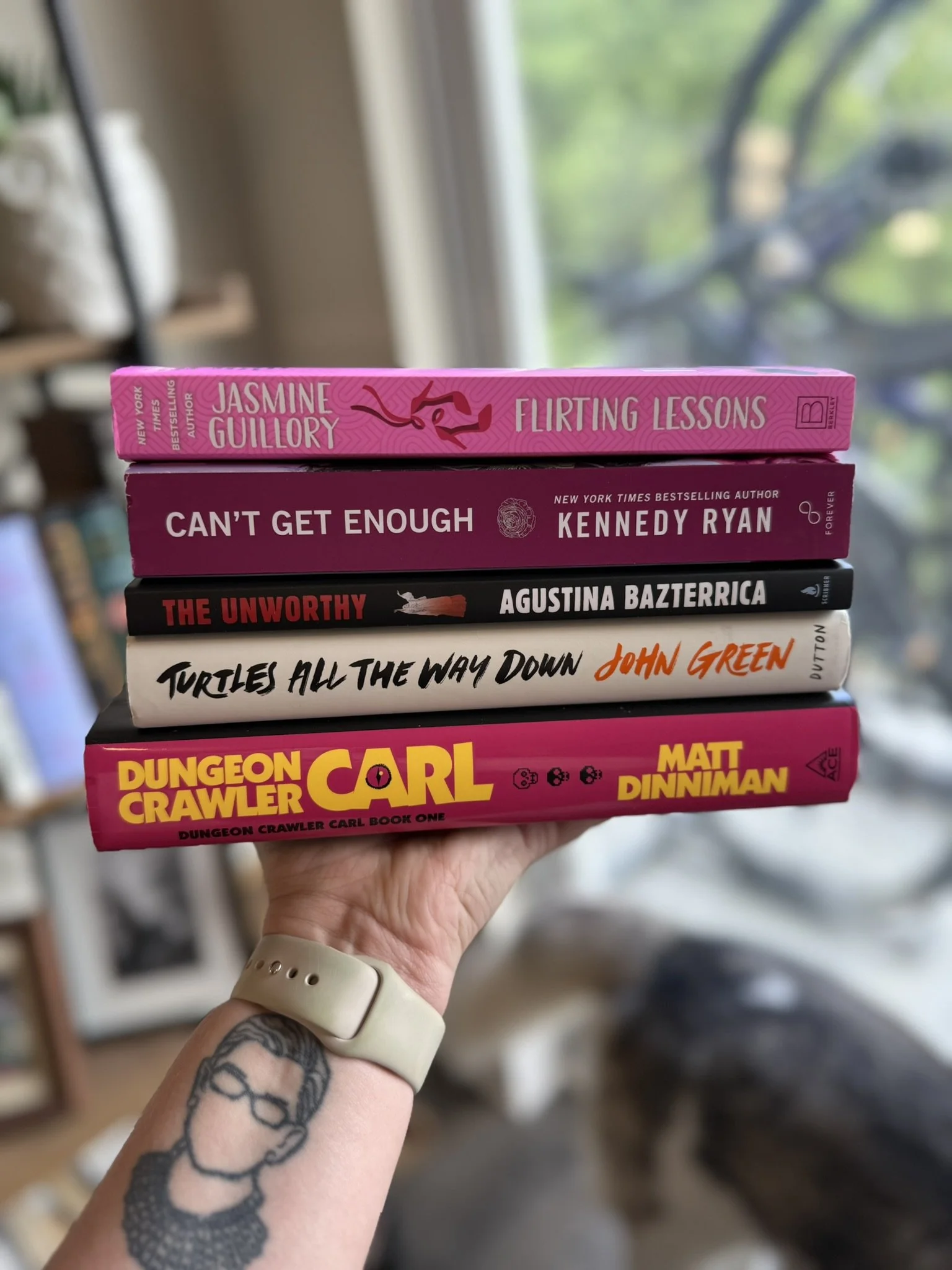 A person with a tattoo on their arm holding a stack of six books. The titles are 'Flirting Lessons' by Jasmine Guillory, 'Can't Get Enough' by Kennedy Ryan, 'The Unworthy' by Agustina Bazterrica, 'Turtles All the Way Down' by John Green, and 'Dungeon