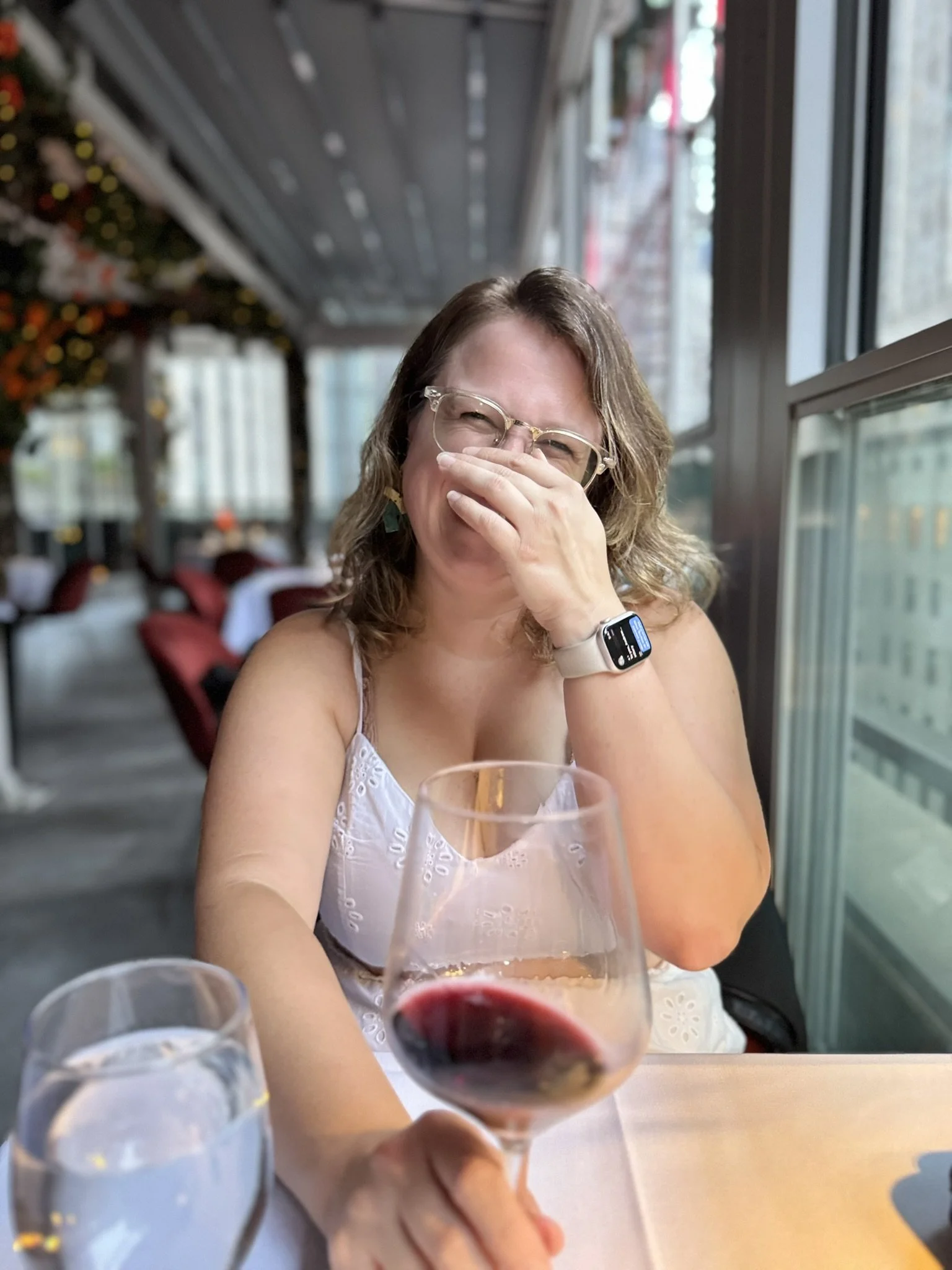 A woman sitting at a restaurant table, smiling and covering her mouth with her hand, with a glass of red wine and a glass of water in the foreground, and decorated restaurant interior in the background.