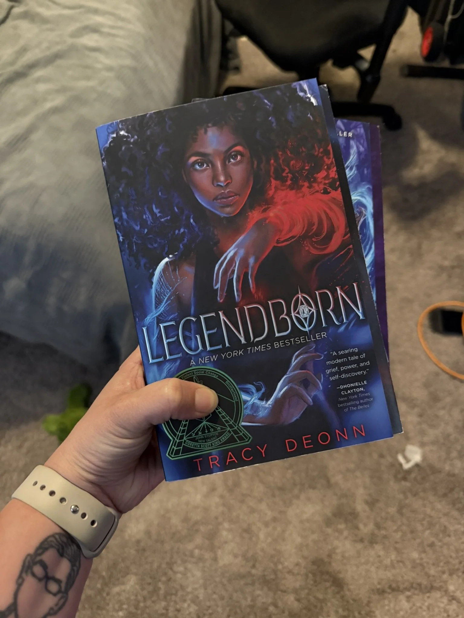 Hand holding a copy of the book 'Legendborn' by Tracy Deonn in a room with carpeted floor and a grey couch.
