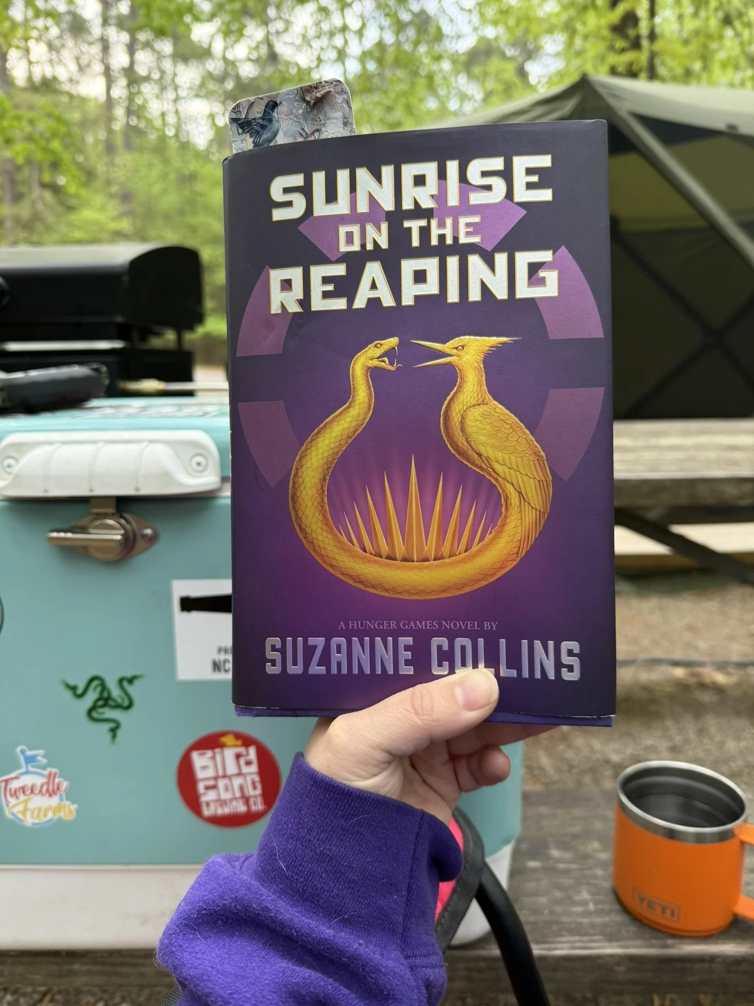 A person holding a copy of the book "Sunrise on the Reaping" by Suzanne Collins outdoors with trees, a picnic table, a camping stove, a sticker, and a cup in the background.