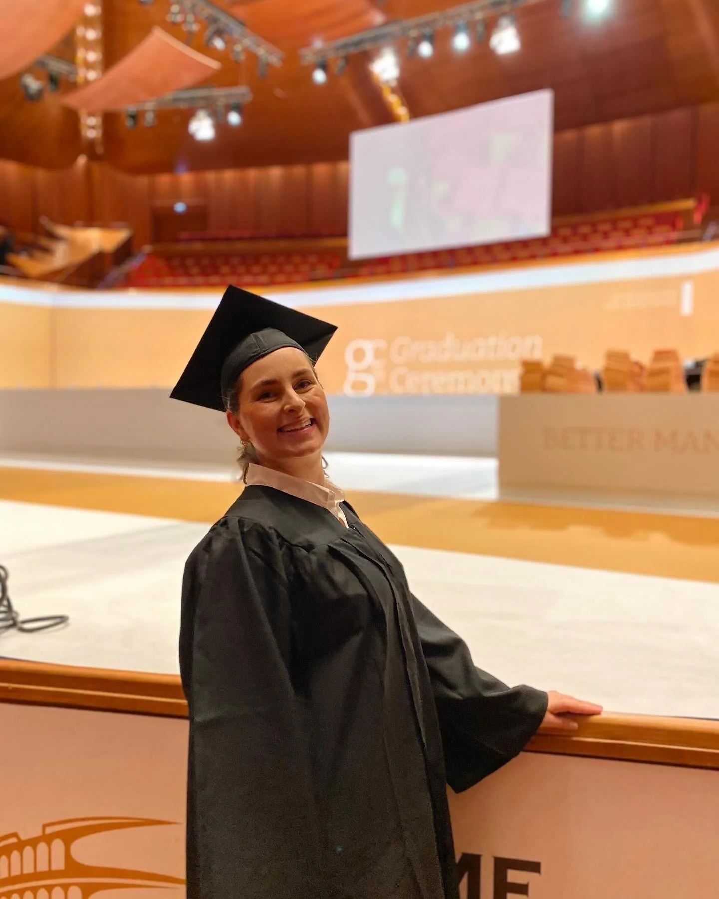 Graduation woman in cap and gown smiling in an auditorium with wooden ceiling and stage in background.