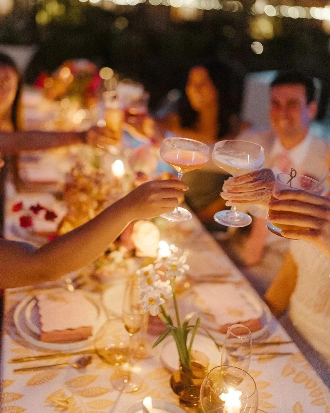 People celebrating around a dinner table with drinks and desserts at an outdoor event during evening or night.