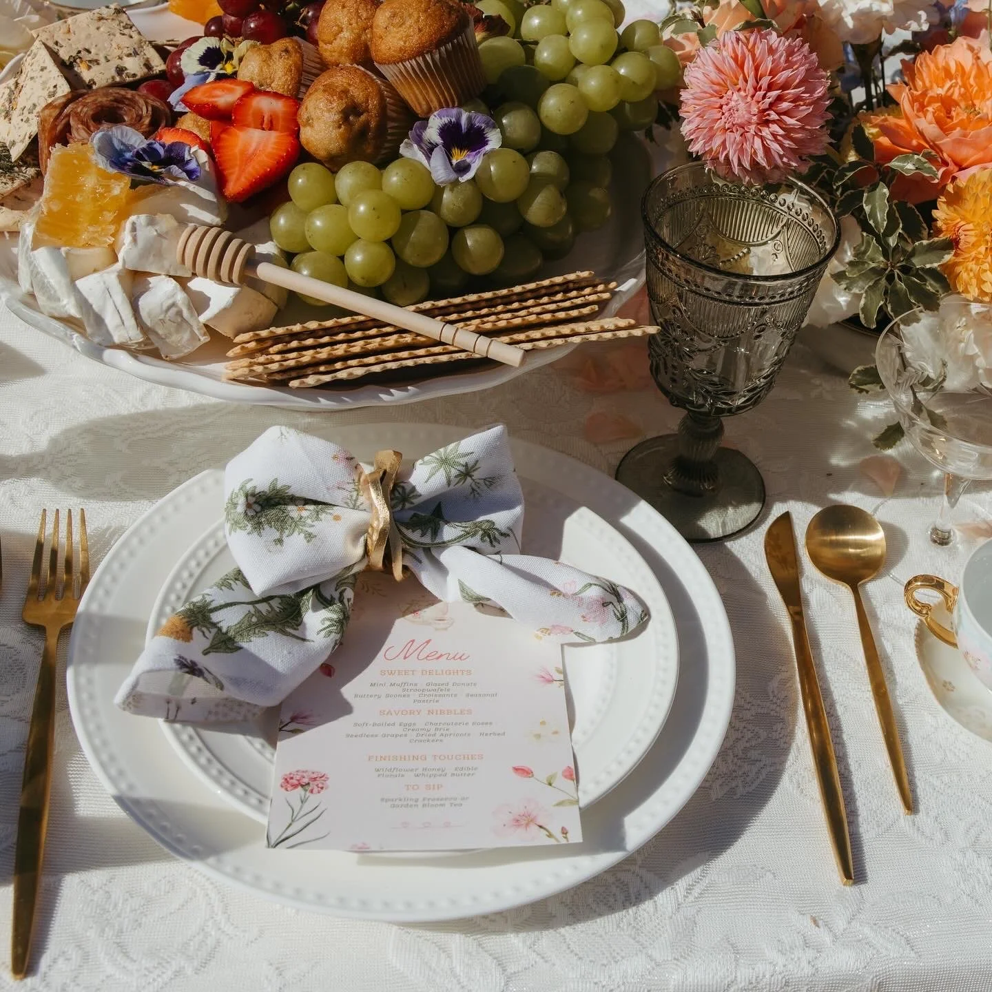 A table setting with a white plate, a menu, a floral napkin, gold flatware, and a glass of water. On the table, there's a large platter with cheese, grapes, strawberries, muffins, crackers, and a honey dipper. Pink and orange flowers decorate the arrangement.