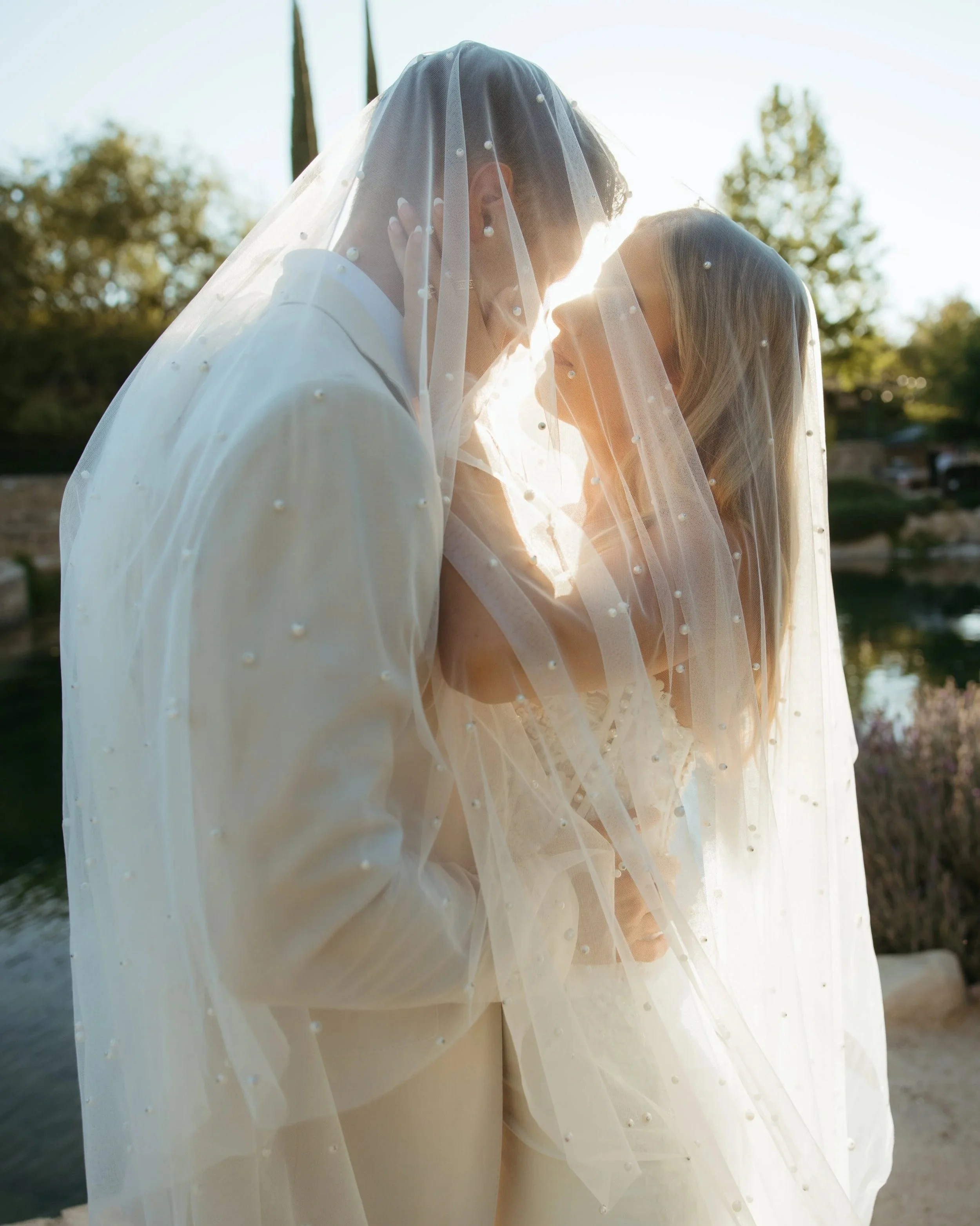 A couple dressed in wedding attire sharing an intimate moment outdoors, with a veil covering both of them, backlit by the sun with trees in the background.