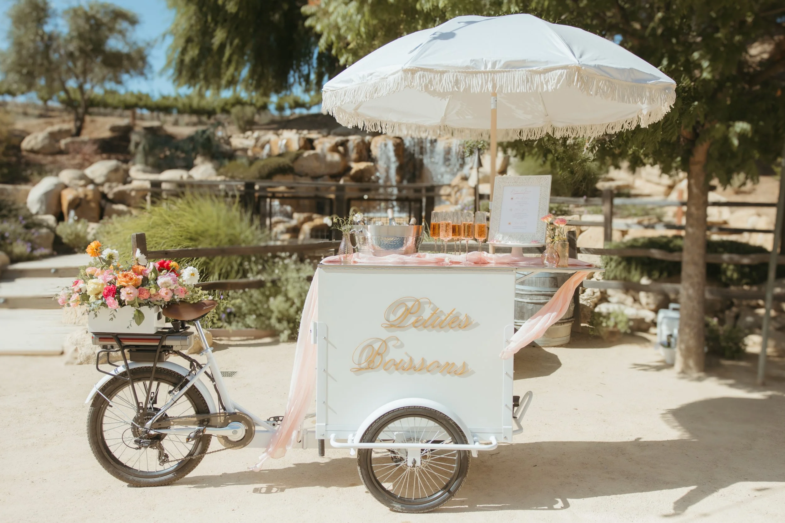 A white tricycle cart with the sign 'Petites Bausons' is set up outdoors under a white umbrella. The cart is decorated with pink ribbons, and holds glasses of pink drinks, a framed menu, and vases with pink and white flowers. In the background, a scenic landscape includes a waterfall, rocks, trees, and a fence.
