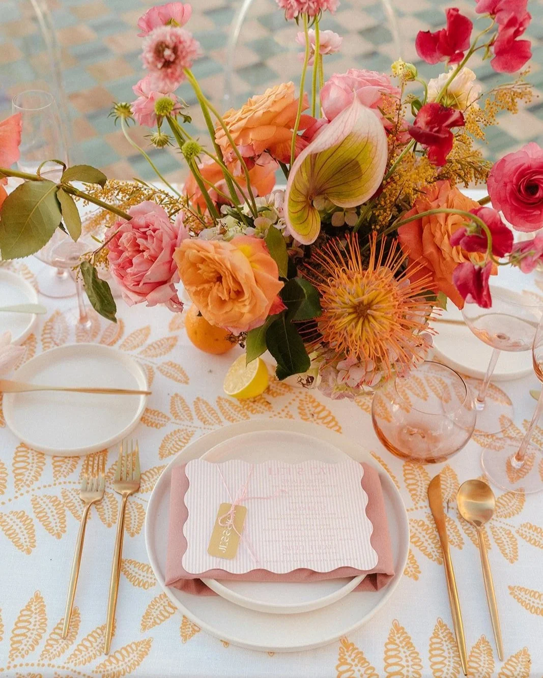 Elegant table setting featuring a large floral centerpiece with pink, peach, and yellow flowers, including roses, anthuriums, and lilies. The table has white and gold cutlery, pink transparent glasses, a white plate with a pink napkin, and a note card. The background shows a patterned tablecloth and a brick floor.
