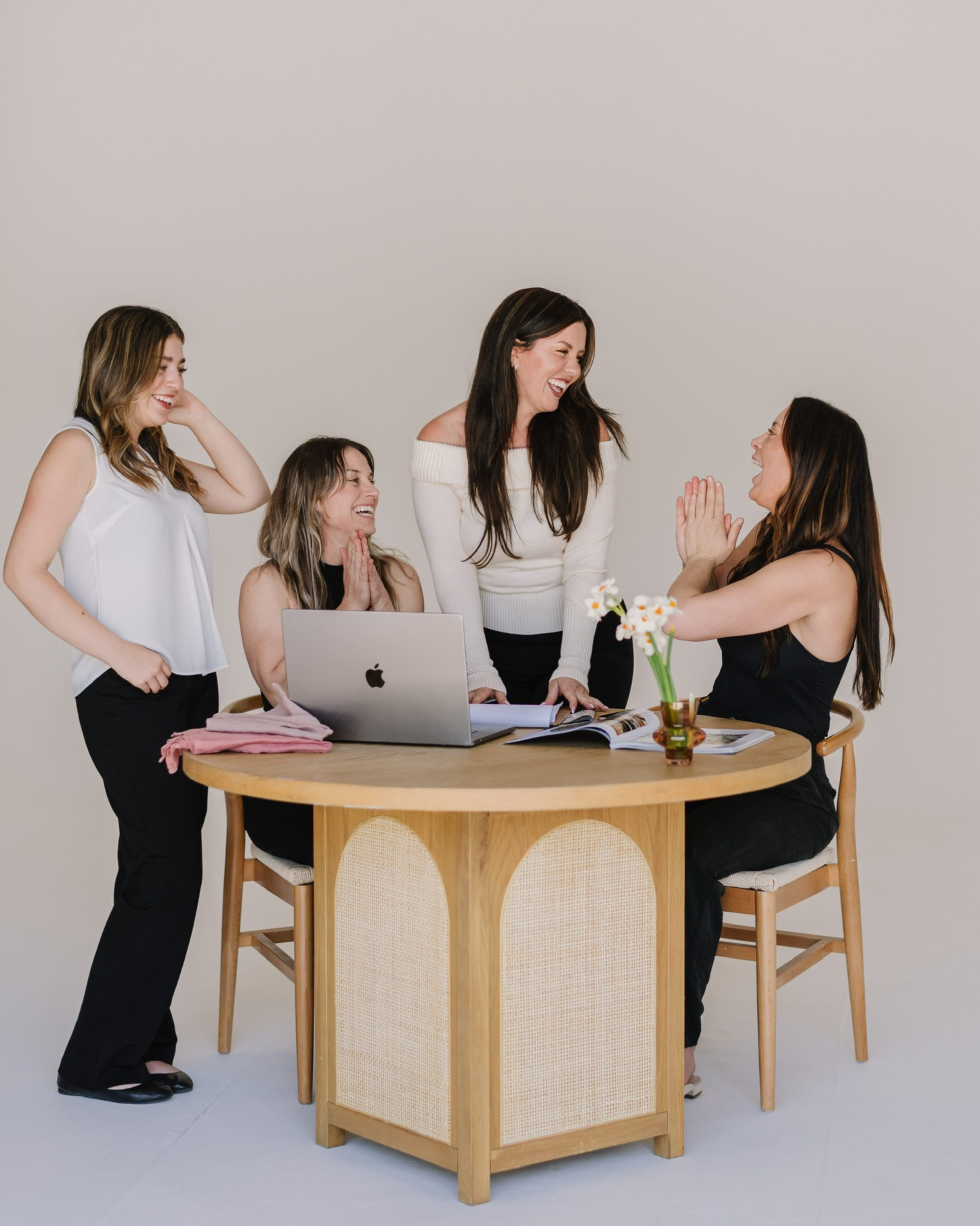 Five women gathered around a round table, smiling and laughing. One woman is sitting at the table with clasped hands, another is standing with her hands on the table, and three are standing nearby, engaging in conversation and smiling. The table has a laptop, books, papers, and a small vase of flowers. The background is plain and light-colored.
